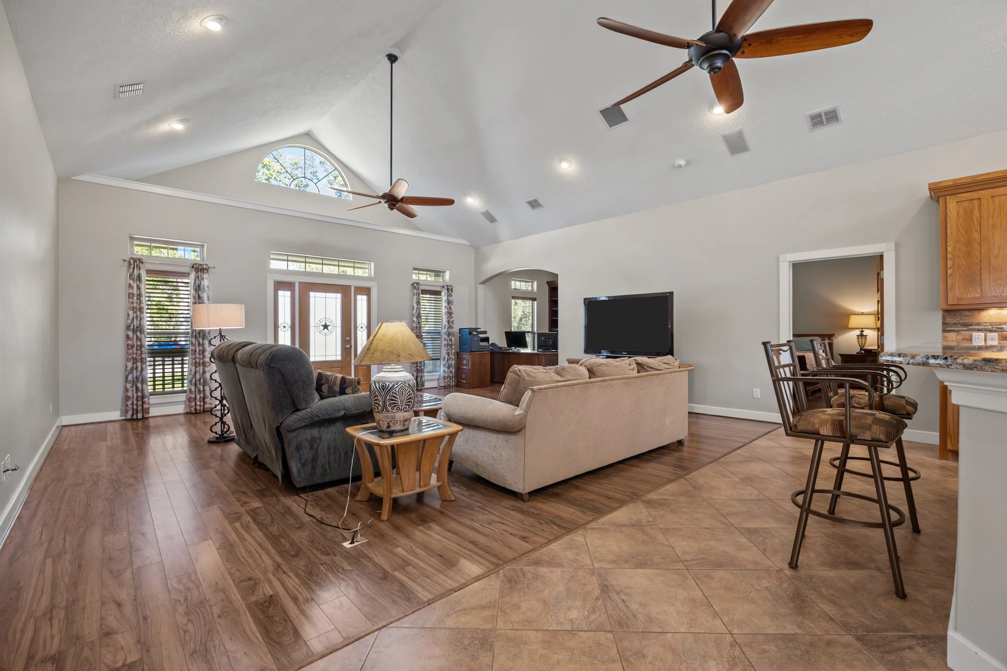 Bright living room with high vaulted ceiling, ceiling fans, hardwood and tiled flooring, sofa, armchair, coffee table with lamp, flat-screen TV, and front door with decorative glass panels.