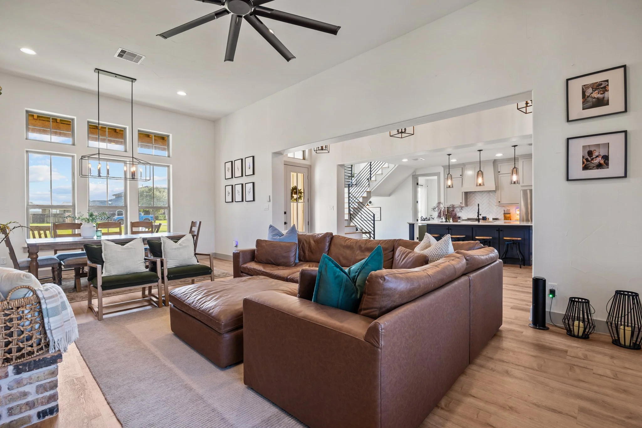 A spacious living room with large windows, a brown leather sectional sofa with teal and beige pillows, a wooden dining table with six chairs near the window, and an open kitchen in the background with pendant lights and white cabinetry. The ceiling has a black modern fan, and there are framed pictures on the walls.