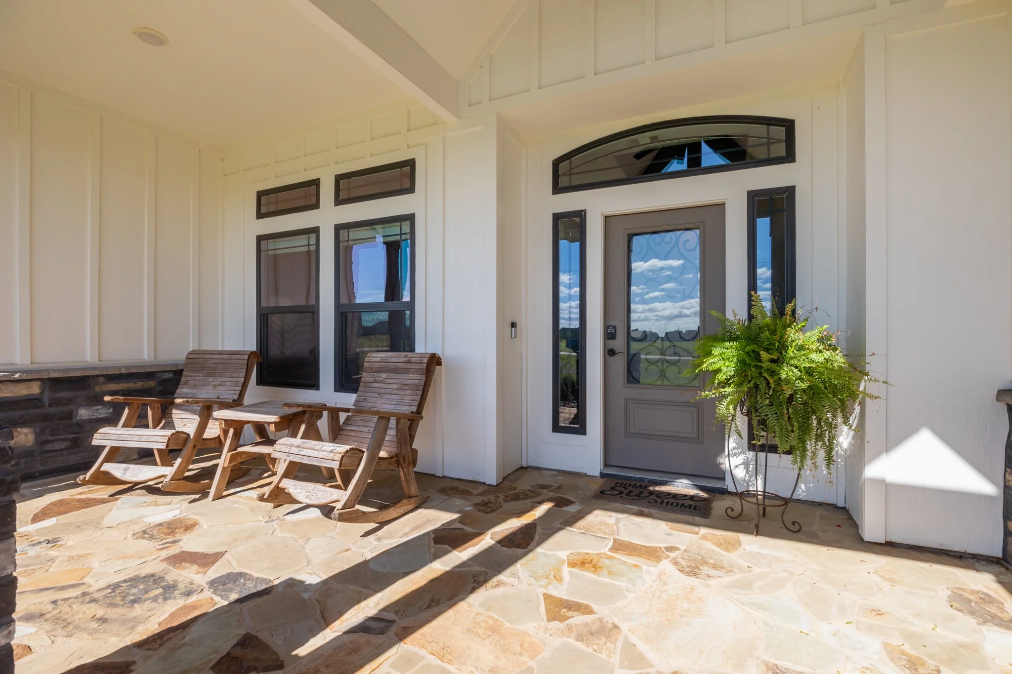 Front porch with two wooden rocking chairs, potted fern, and a door with windows and a glass panel, under a covered roof, with a stone floor.
