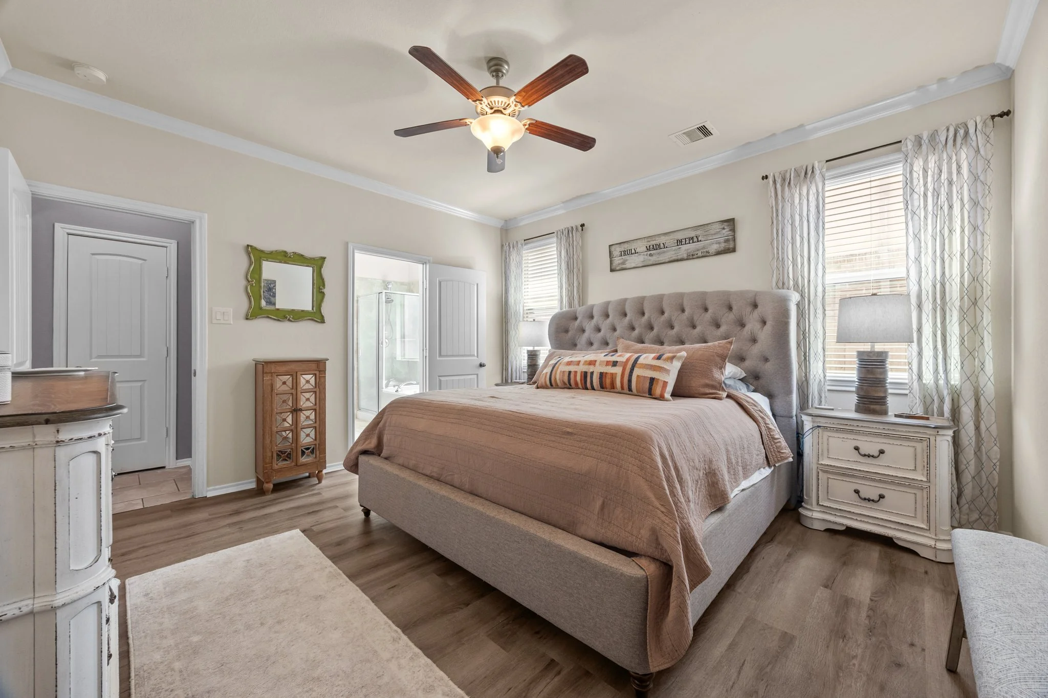 A bedroom with a large bed, beige bedding, tufted headboard, two nightstands with lamps, windows with patterned curtains, a ceiling fan, and a door leading to a bathroom with a shower.