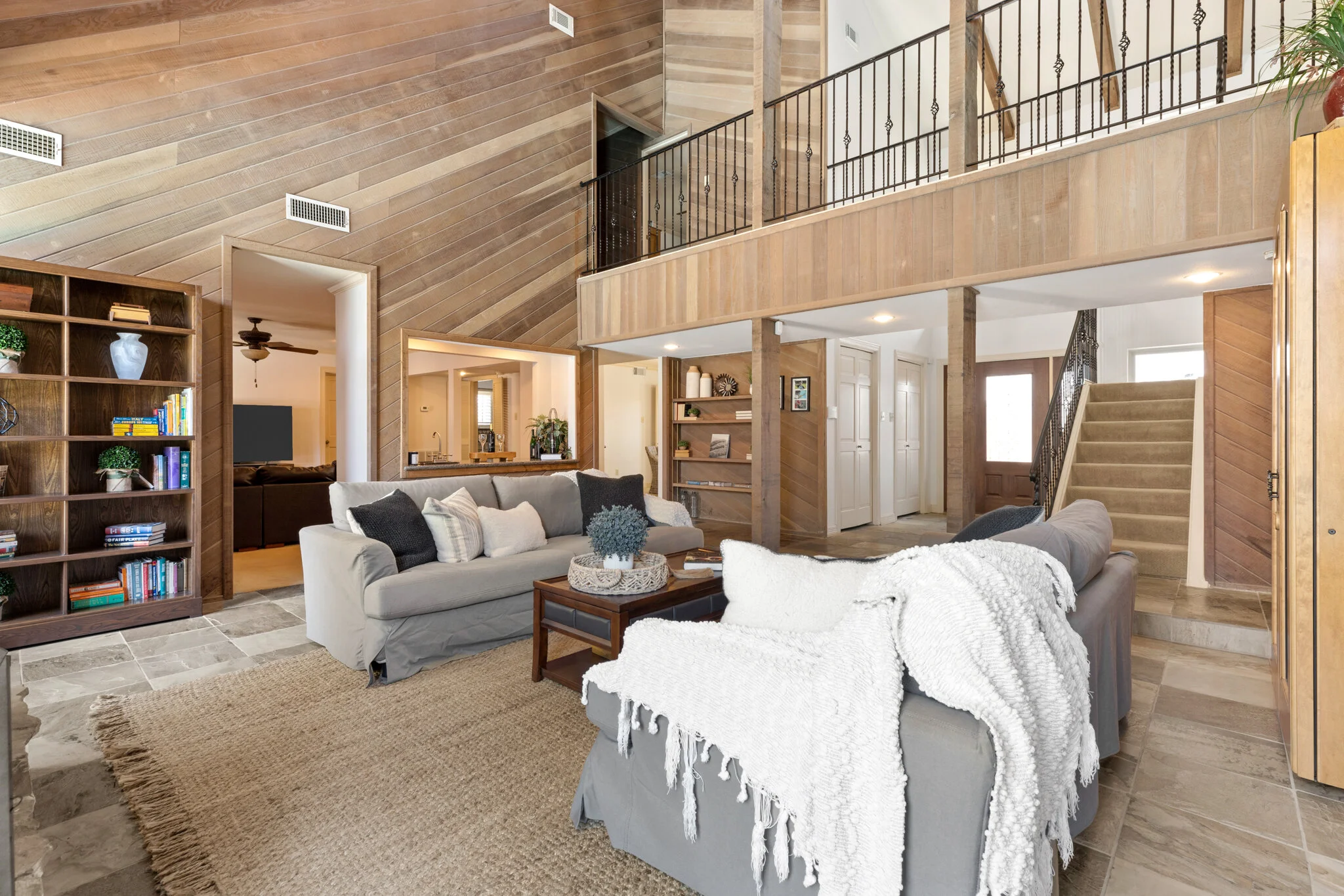 Living room with beige sofas, a wooden bookshelf, a textured rug, and wooden wall paneling in a two-story home with an open loft area and stairs leading to the entrance.