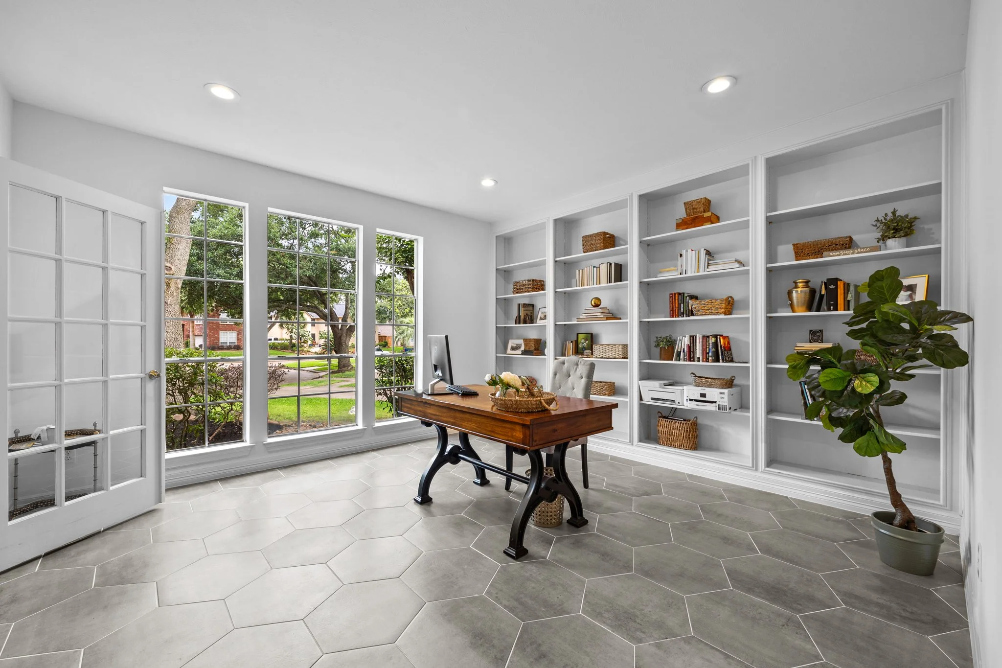 Bright home office with large windows, a wooden desk, a computer, a beige cushioned chair, built-in white bookshelves with books and decorative baskets, a potted plant, and a view of a green yard outside.
