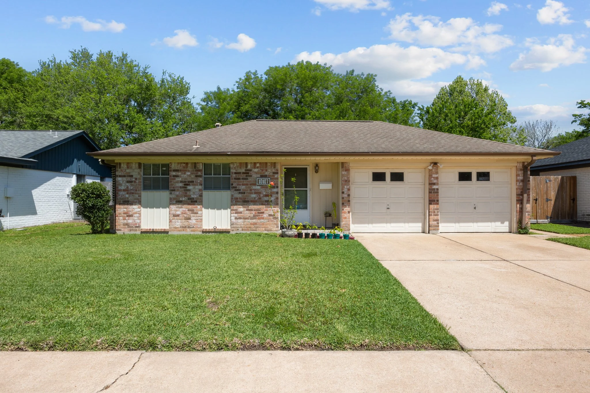 A single-story suburban house with brick and siding exterior, a two-car garage, a manicured front lawn, and a concrete driveway. The house has windows with awnings and a small front porch with potted plants. Tall green trees are in the background under a partly cloudy sky.