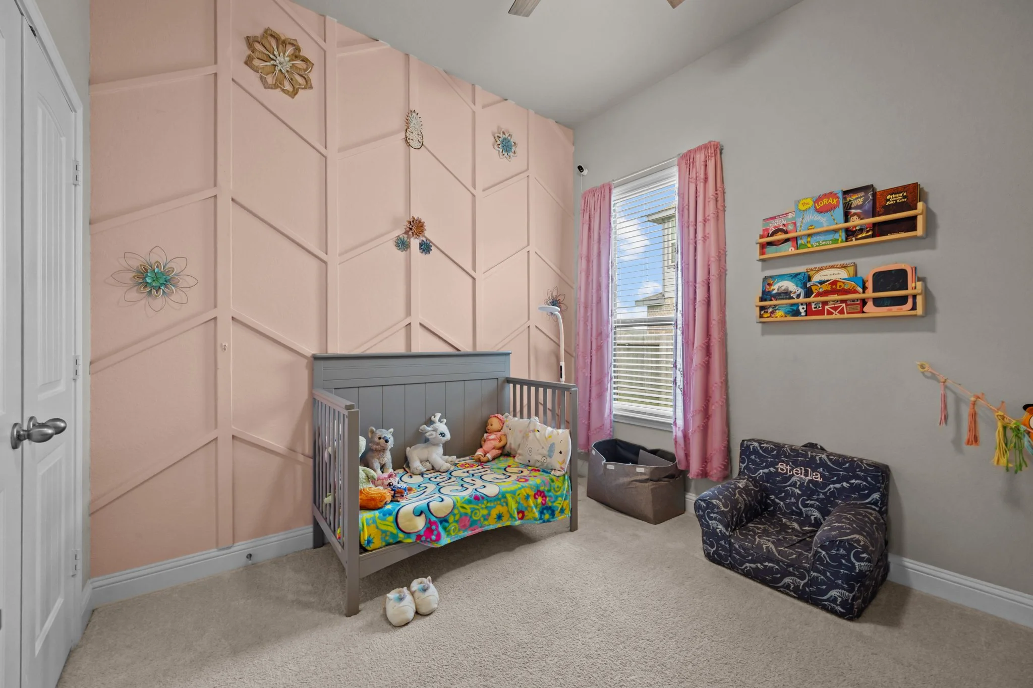 A children's bedroom with pink wall decoration, plush toys in a crib, a small patterned armchair with the name Stella embroidered, a window with pink curtains, and shelves with children's books.