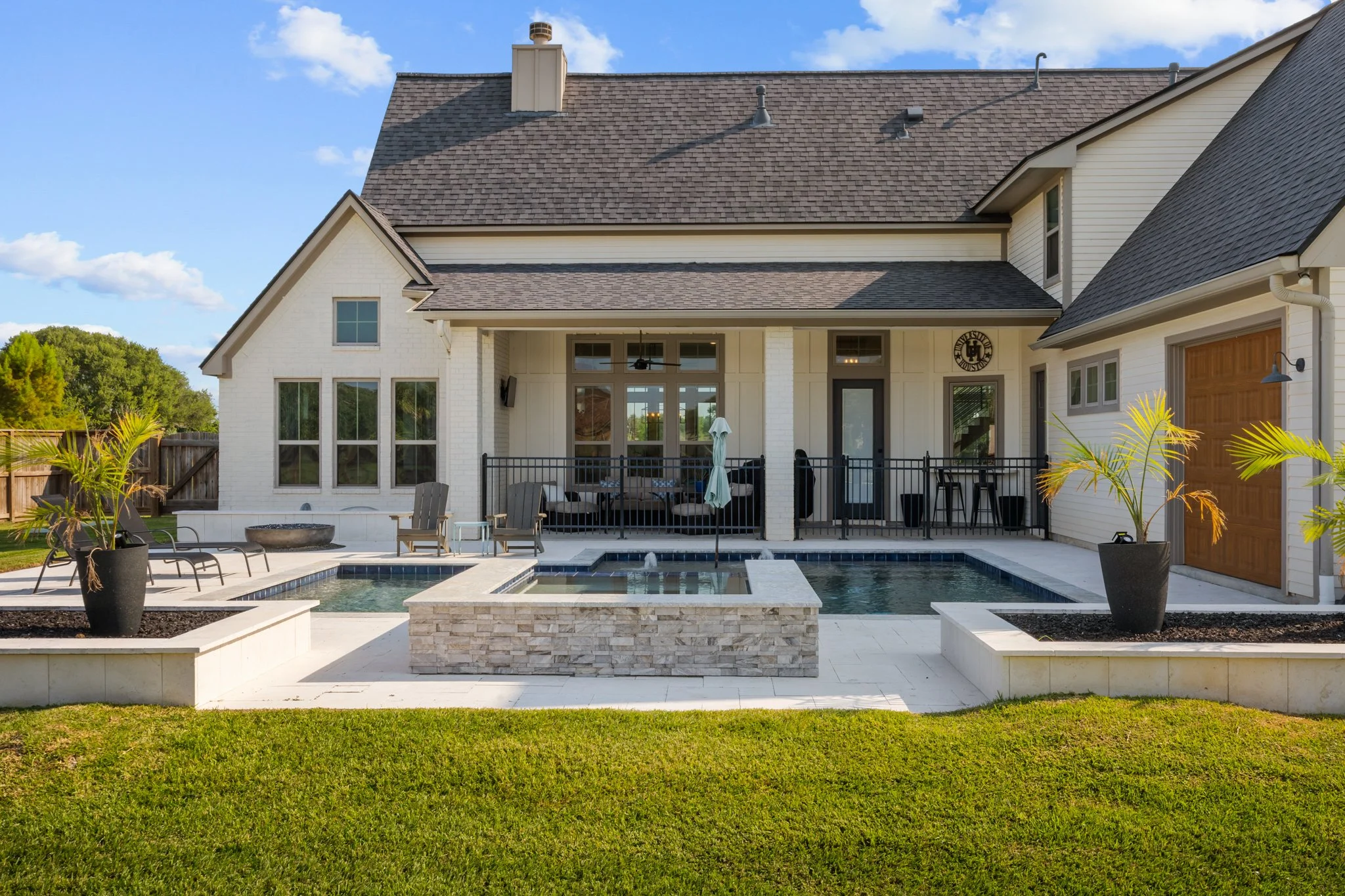  Backyard view of a modern house with a swimming pool, hot tub, potted plants, lawn, and patio furniture under a blue sky.
