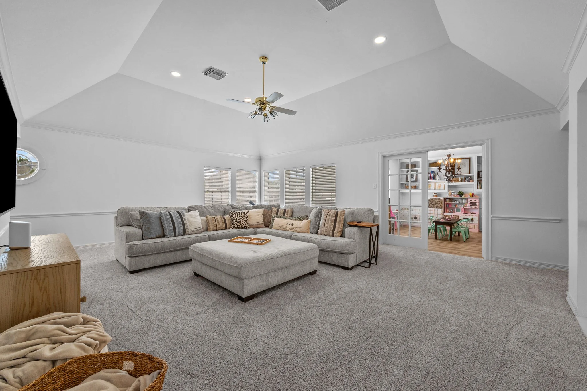 Spacious, white living room with a high, vaulted ceiling, ceiling fan, multiple windows with blinds, a large gray sectional sofa with throw pillows, a ottoman in front, a side table, and a view into a decorated playroom with tables, chairs, and bookshelves.