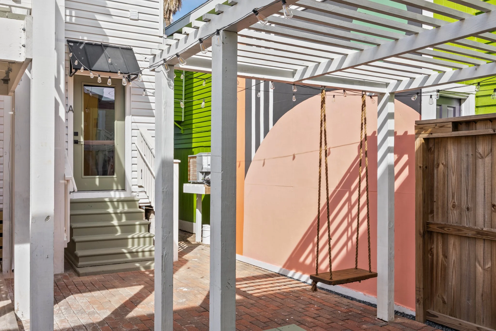 Backyard patio area with a swing hanging from a wooden pergola, string lights, colorful painted walls, and a small outdoor sink.