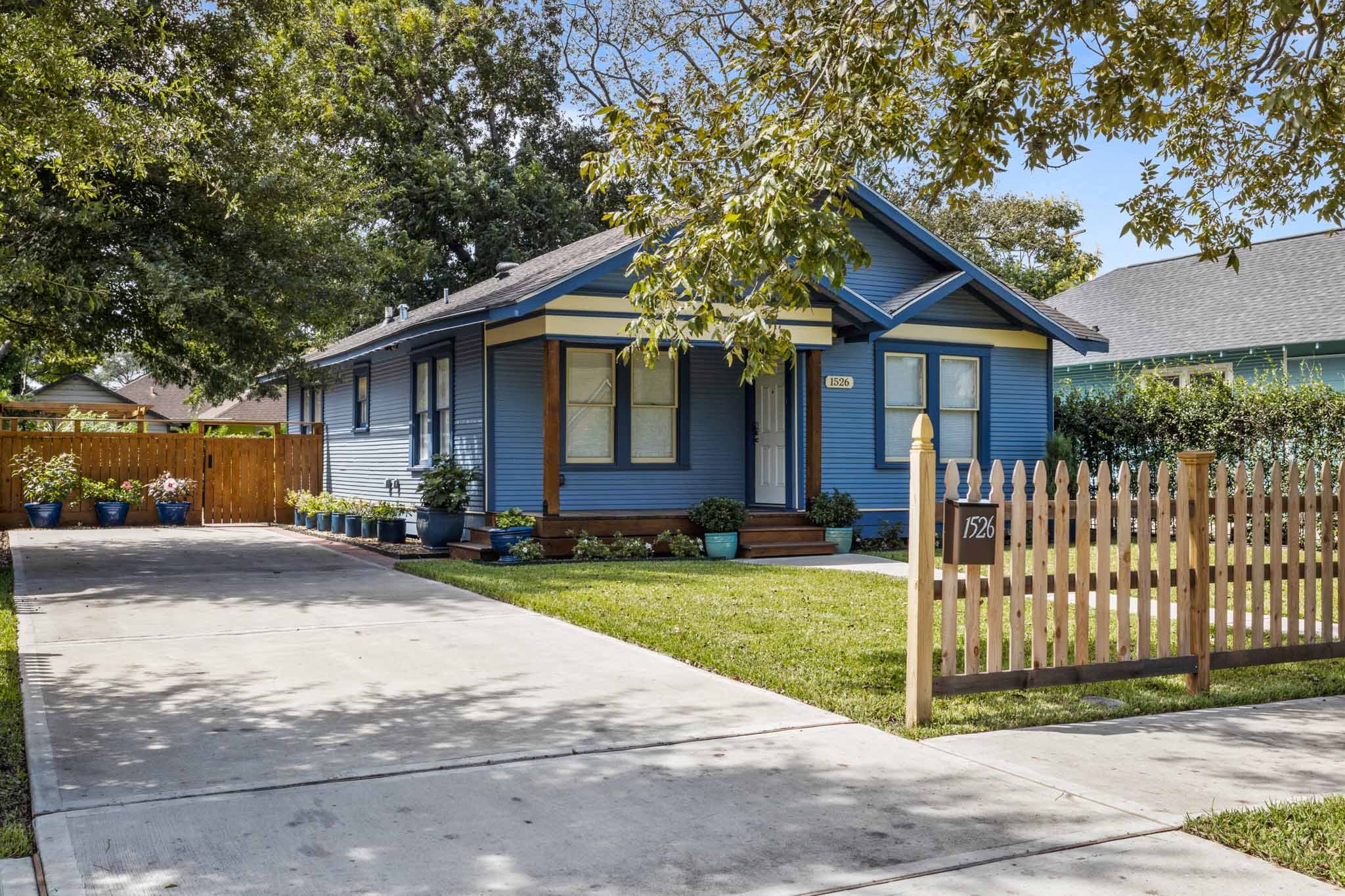A blue house with a small front yard, surrounded by a wooden fence, with potted plants on the porch and along the driveway, under a tree in a suburban neighborhood.
