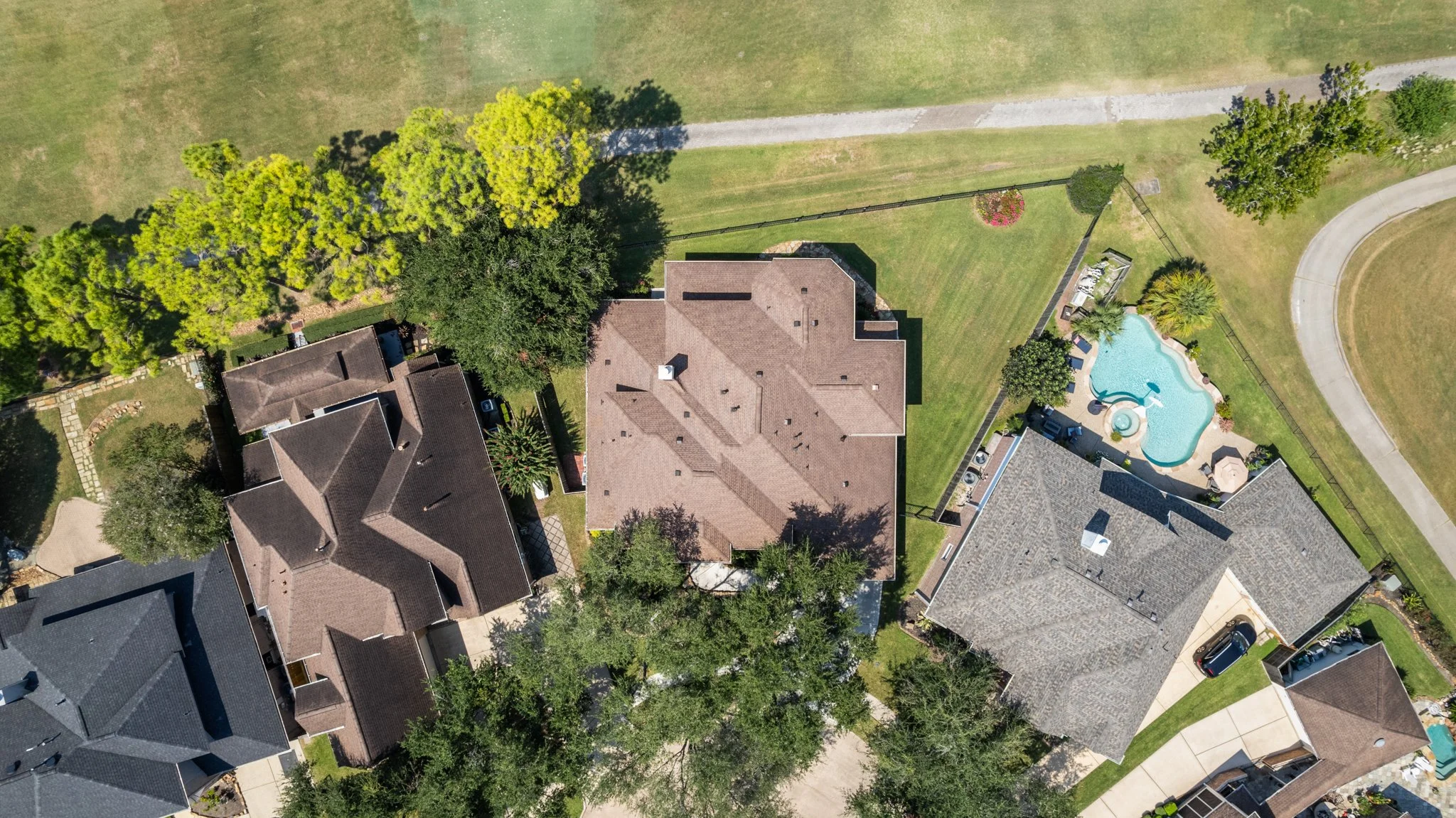 Aerial view of a residential neighborhood with several houses, green lawns, trees, a swimming pool with a hot tub, and paved sidewalks.