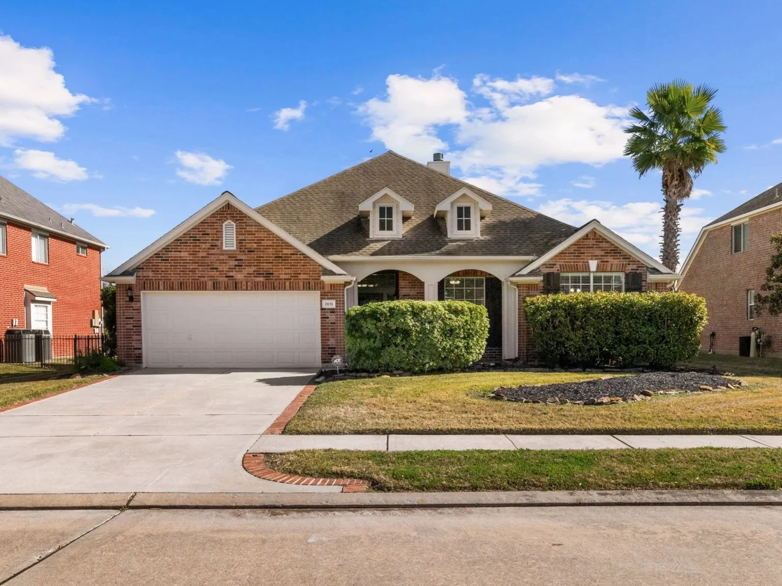 A brick suburban house with a two-car garage, a landscaped front yard, and a palm tree, under a blue sky with scattered clouds.