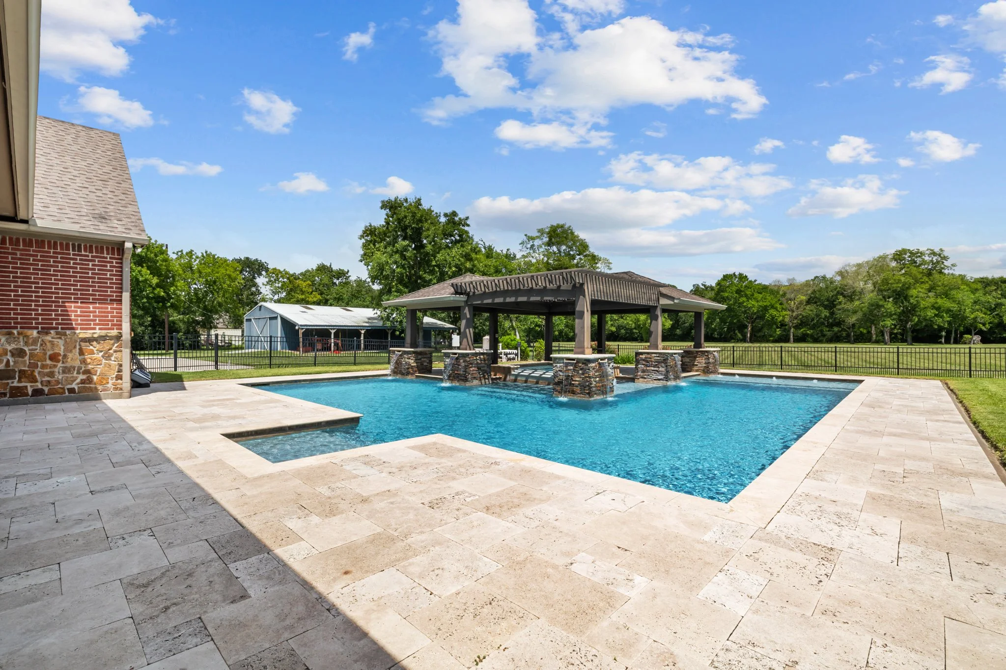 Empty swimming pool with stone deck, shaded pavilion with stone pillars, surrounded by green grass, trees, and fence under a blue sky with clouds.