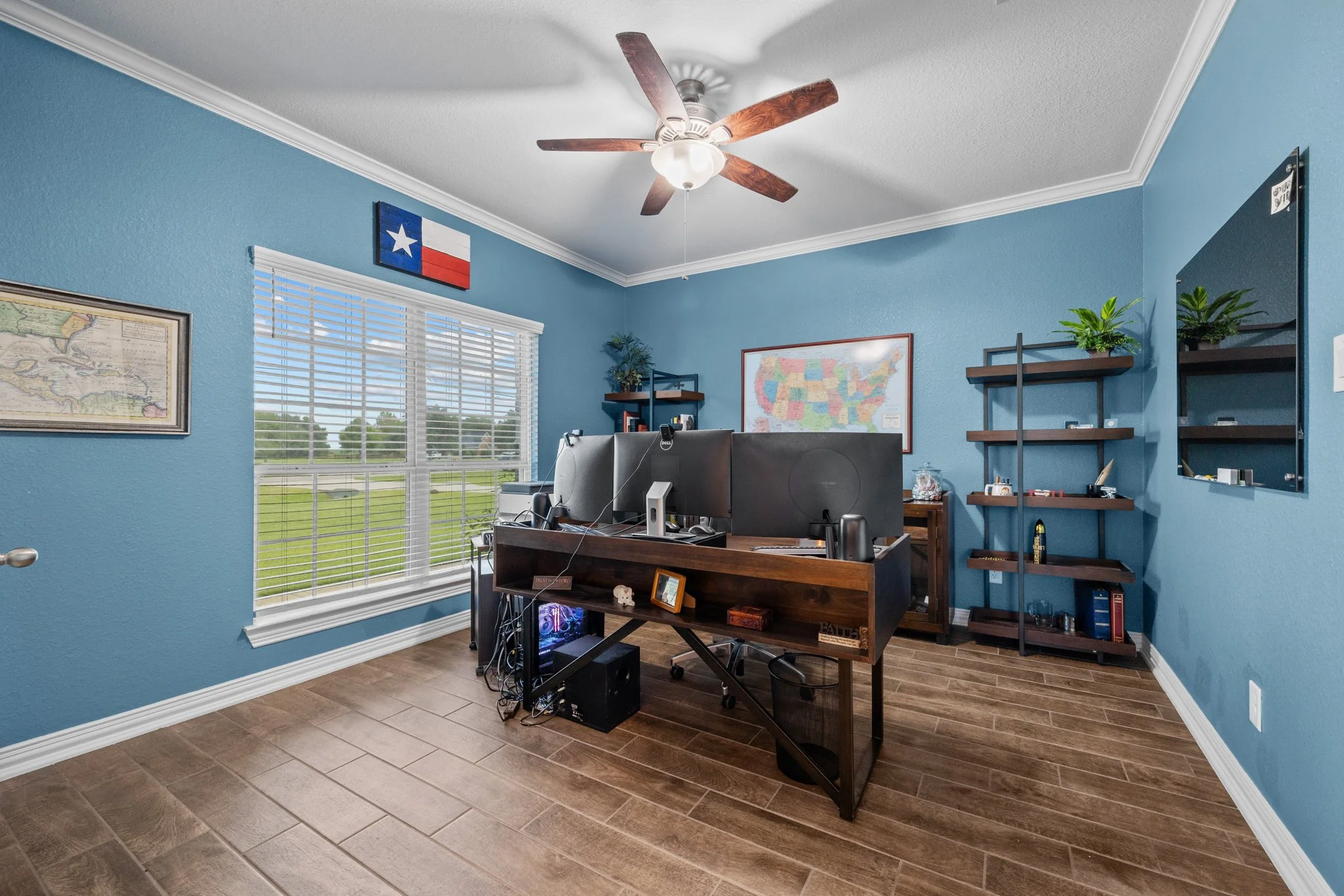 This image shows a home office with blue walls, a large window with blinds, a ceiling fan, and various decorations including a Texas flag. There is a desk with dual monitors, a computer tower underneath, and shelves with plants and books. The floor is wood, and there's a mirror on the wall.