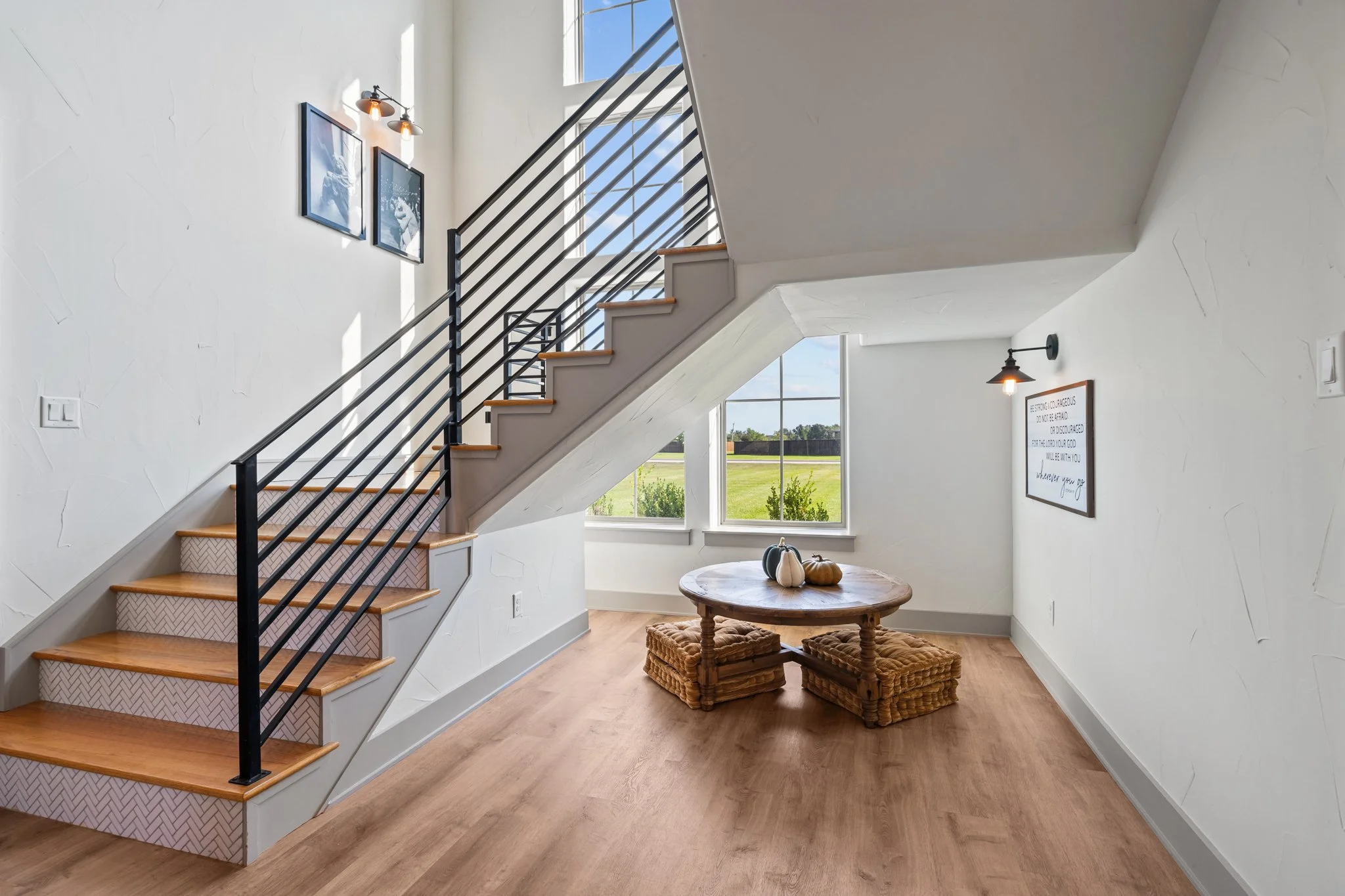 A modern home interior with a staircase leading to an upper level, a round table with pumpkins and gourds, large windows letting in natural light, and minimal decor including framed pictures and wall sconces.