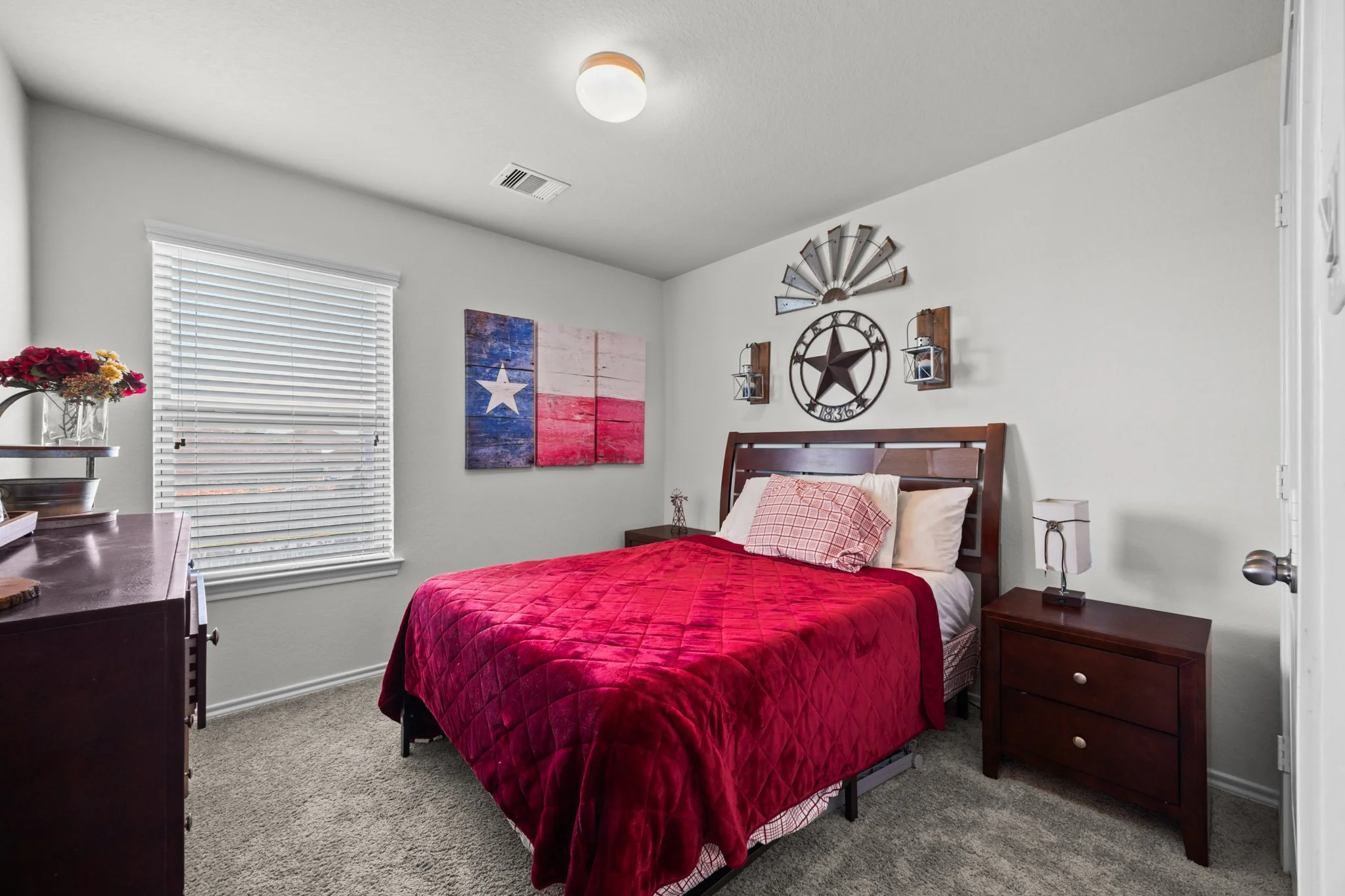 Bedroom with a bed covered in a red quilt and white pillows, dark wood nightstand with a lamp, window with closed blinds, Texas flag artwork on the wall, and decorative Texas-themed wall hangings.