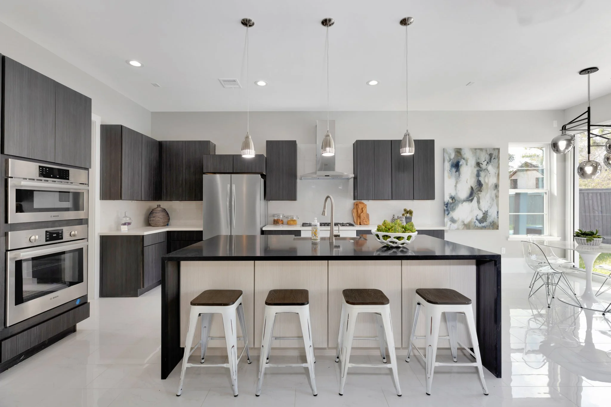 Modern kitchen with dark gray cabinets, stainless steel appliances, black island with white panels and four white bar stools, pendant lighting, and a breakfast nook with a circular table and glass chairs by a window.