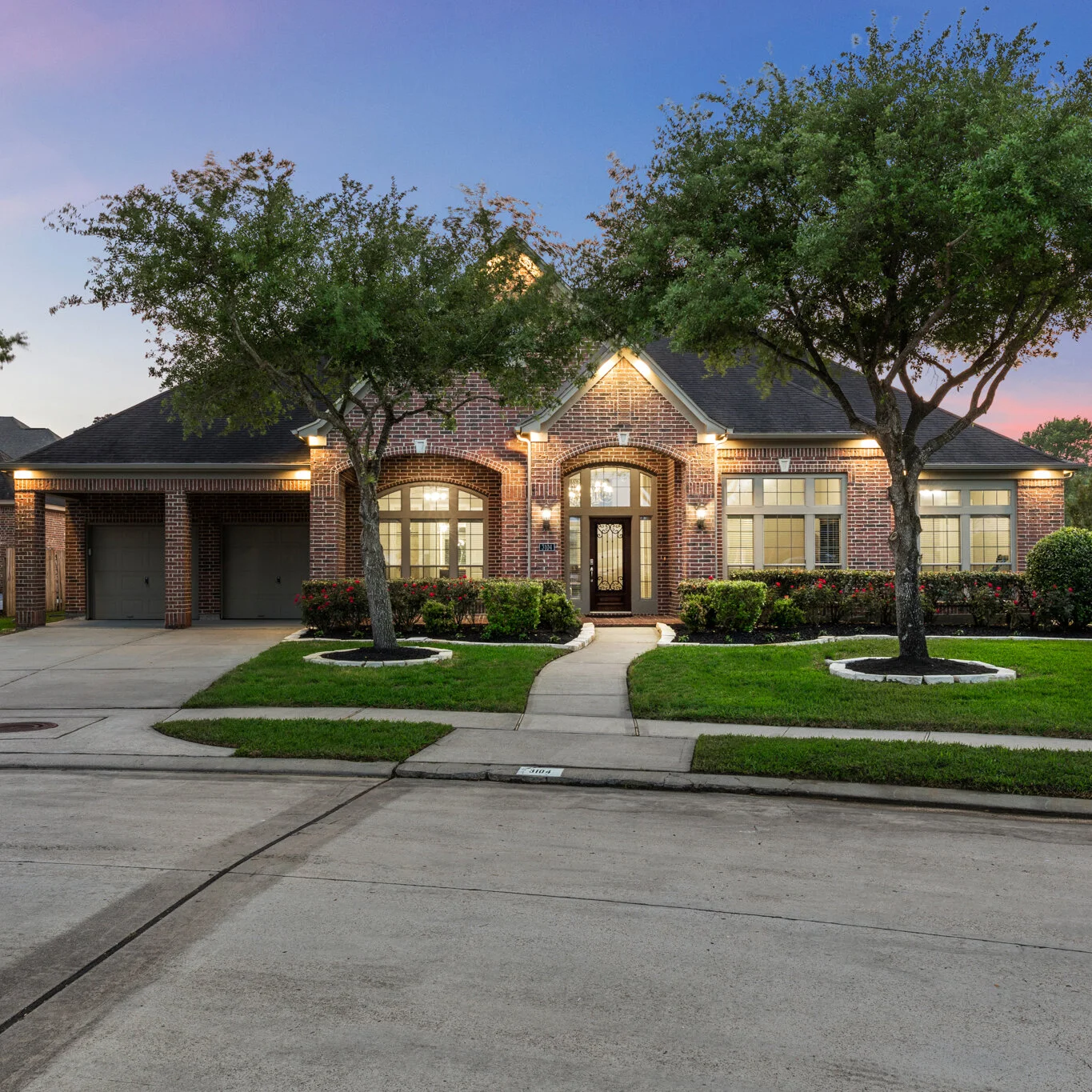 A brick house with a two-car garage, front lawn, and trees, illuminated by exterior lighting at dusk.