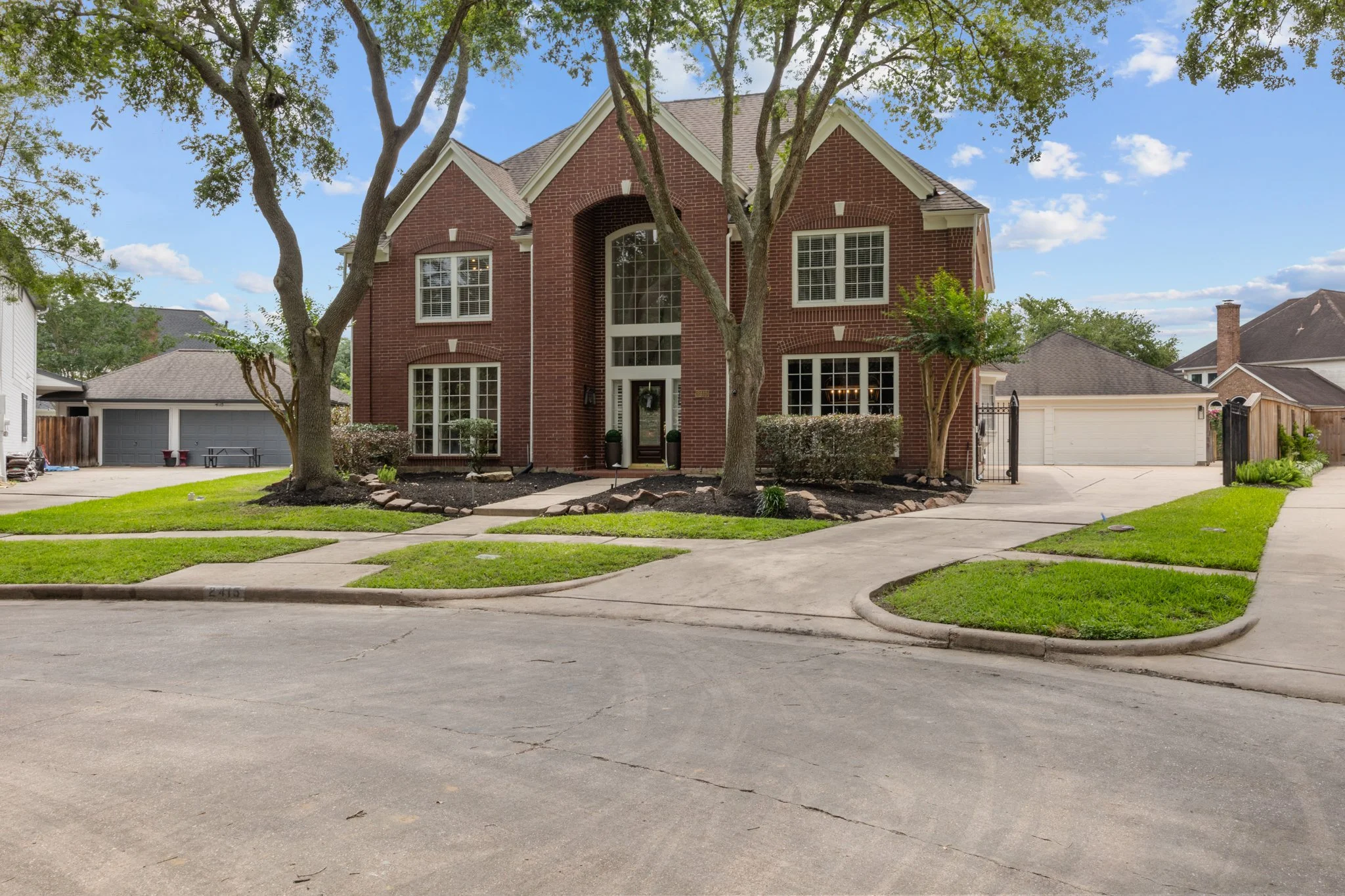 Front view of a two-story brick house with large windows, surrounded by a well-maintained lawn, trees, and a concrete driveway leading to a gated side entrance and garage, under a partly cloudy sky.