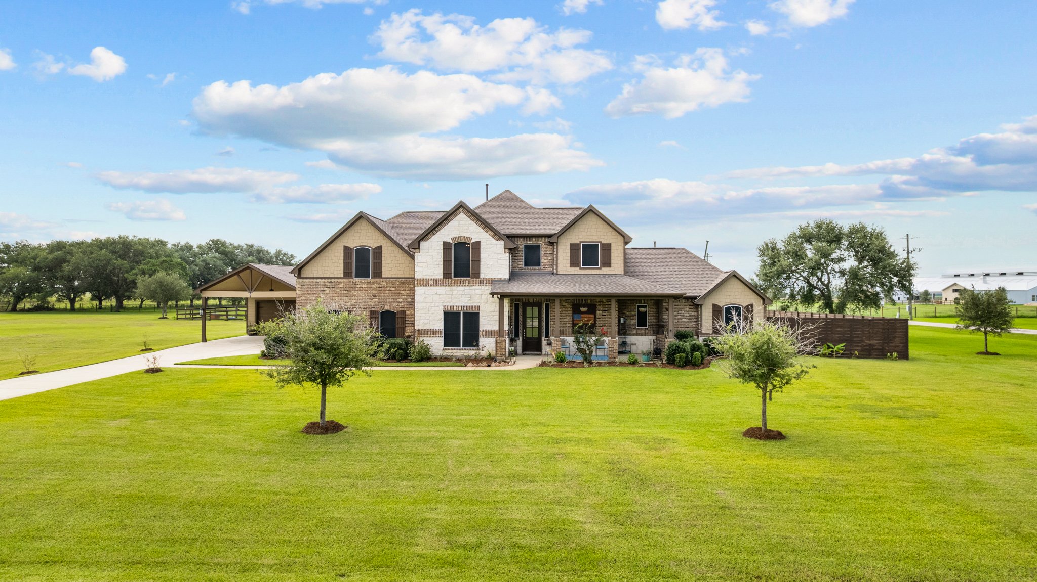 A large two-story house with brick and beige siding, surrounded by a well-maintained lawn with three small trees, under a partly cloudy sky.