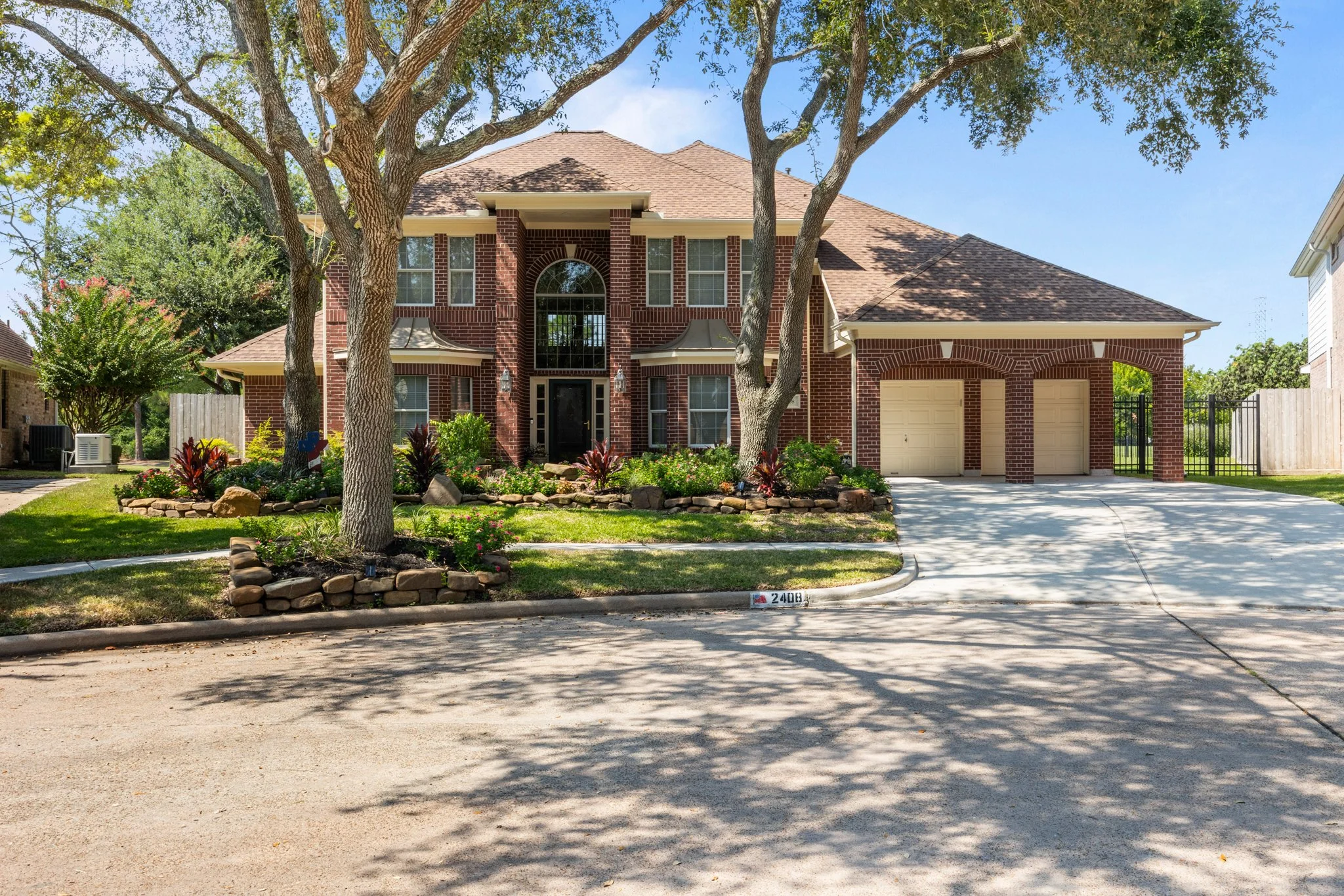 Front view of a large brick house with an attached garage, surrounded by green trees and landscaping with plants and rocks.