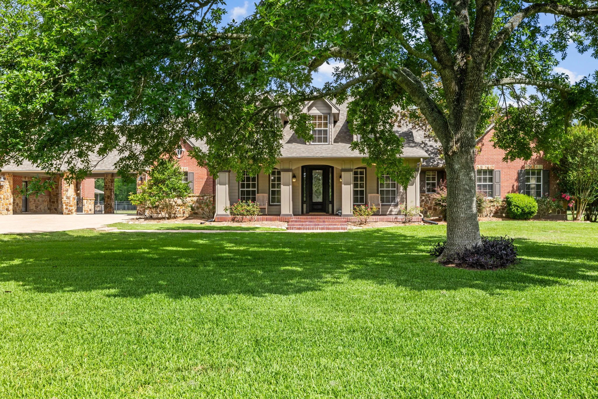 Front view of a large house with brick and gray siding, surrounded by a green lawn and trees, with a paved driveway on the left.