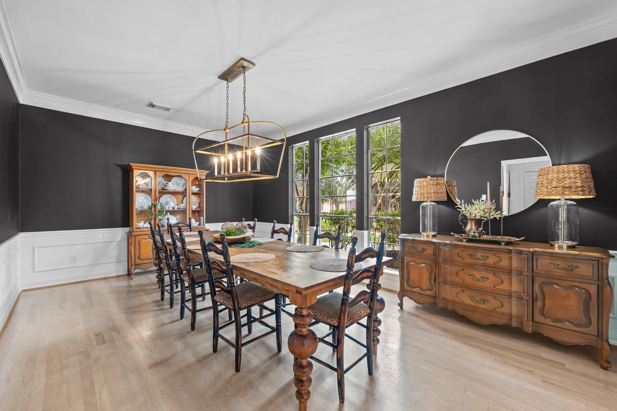 A modern dining room with a wooden table, black chairs, a black accent wall, a wooden sideboard with two lamps and a round mirror, a wooden china cabinet, and large windows showing greenery outside.