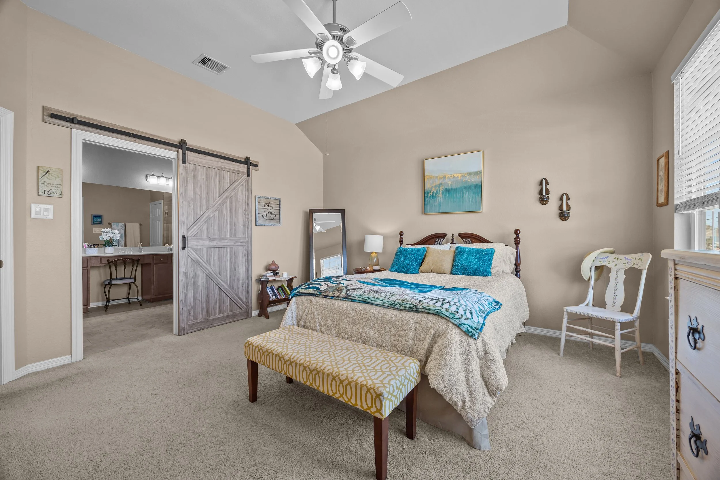 A bedroom with a bed that has a beige bedspread and blue pillows, a yellow and white patterned bench at the foot of the bed, a white chair near the window, and a wooden dresser. There's a ceiling fan and a wall art above the bed. A sliding barn door leads to a bathroom area.