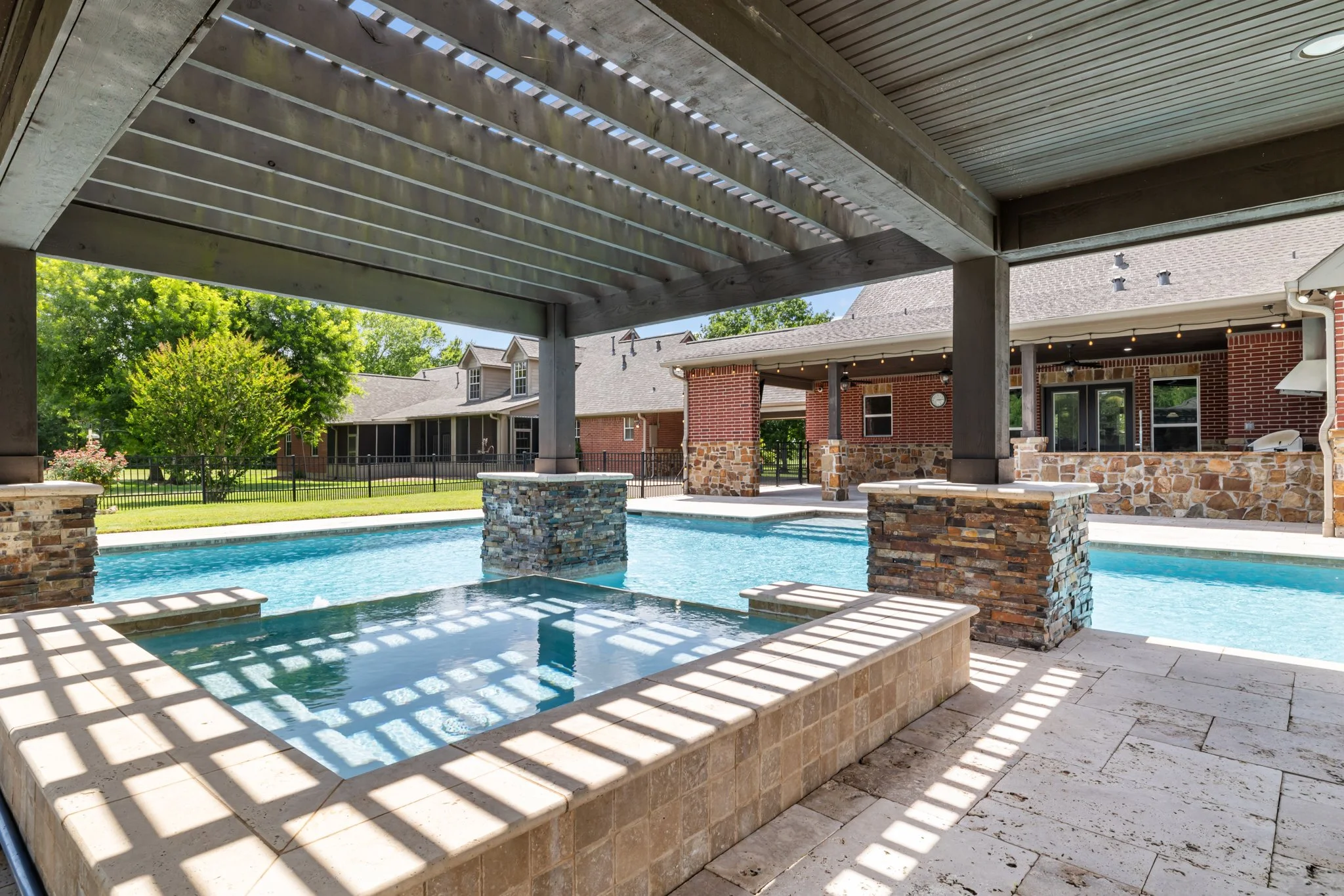 View of a backyard pool area with a shaded patio, brick and stone house, and green trees in the background.
