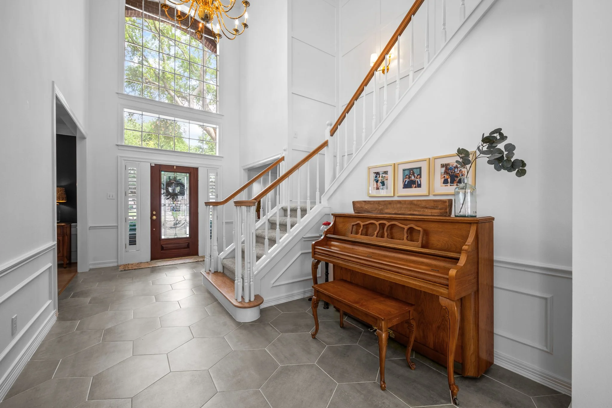 Entrance foyer of a house featuring a wooden front door with glass panels, a staircase with a wooden handrail, a vintage upright piano, pictures hanging on the wall, large windows with greenery outside, and a chandelier hanging from the ceiling.