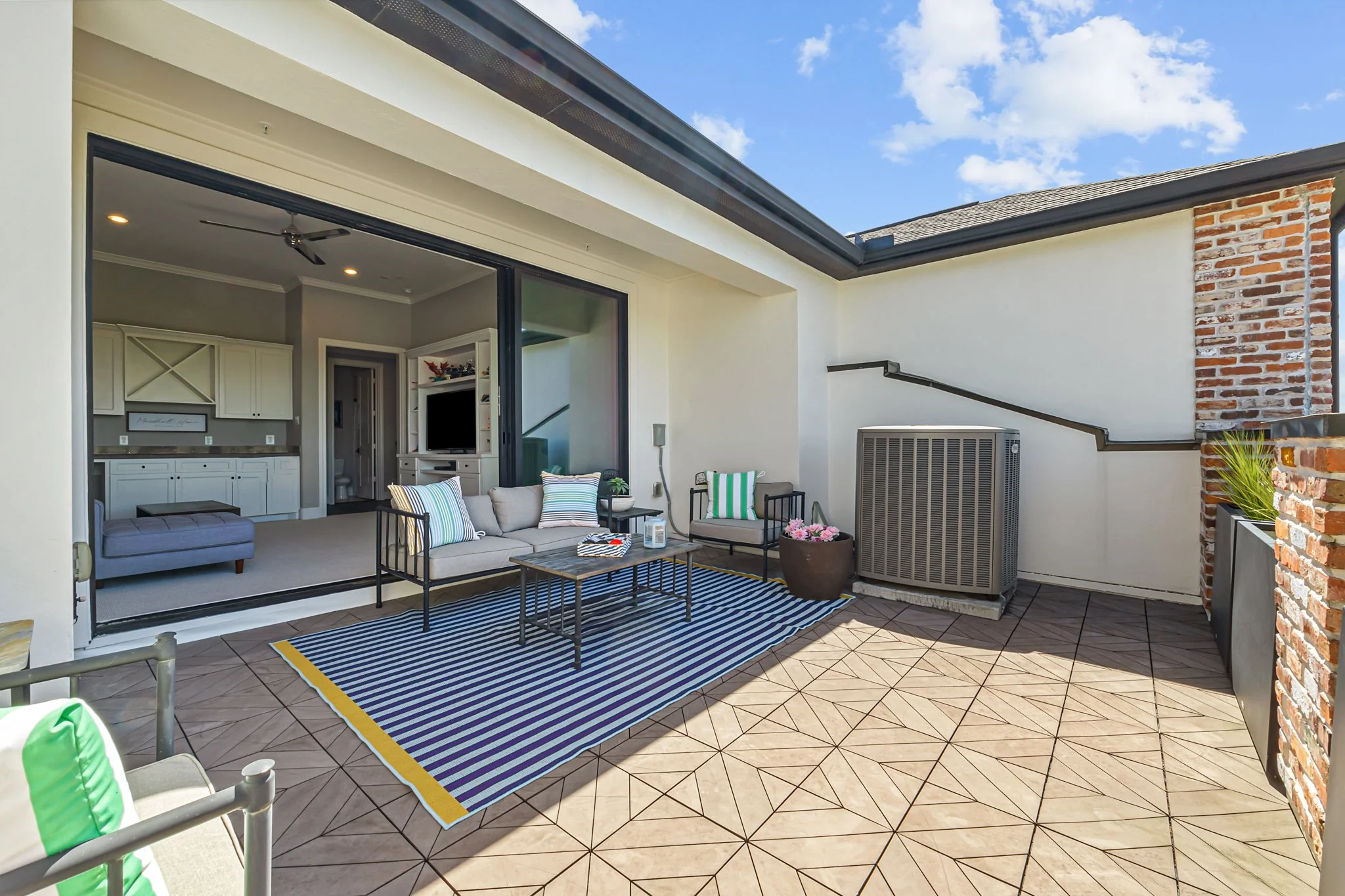 Outdoor balcony with tiled flooring, striped rug, coffee table, and lounge chairs with striped cushions, overlooking an open sliding glass door leading to a living room with built-in cabinets and a TV.
