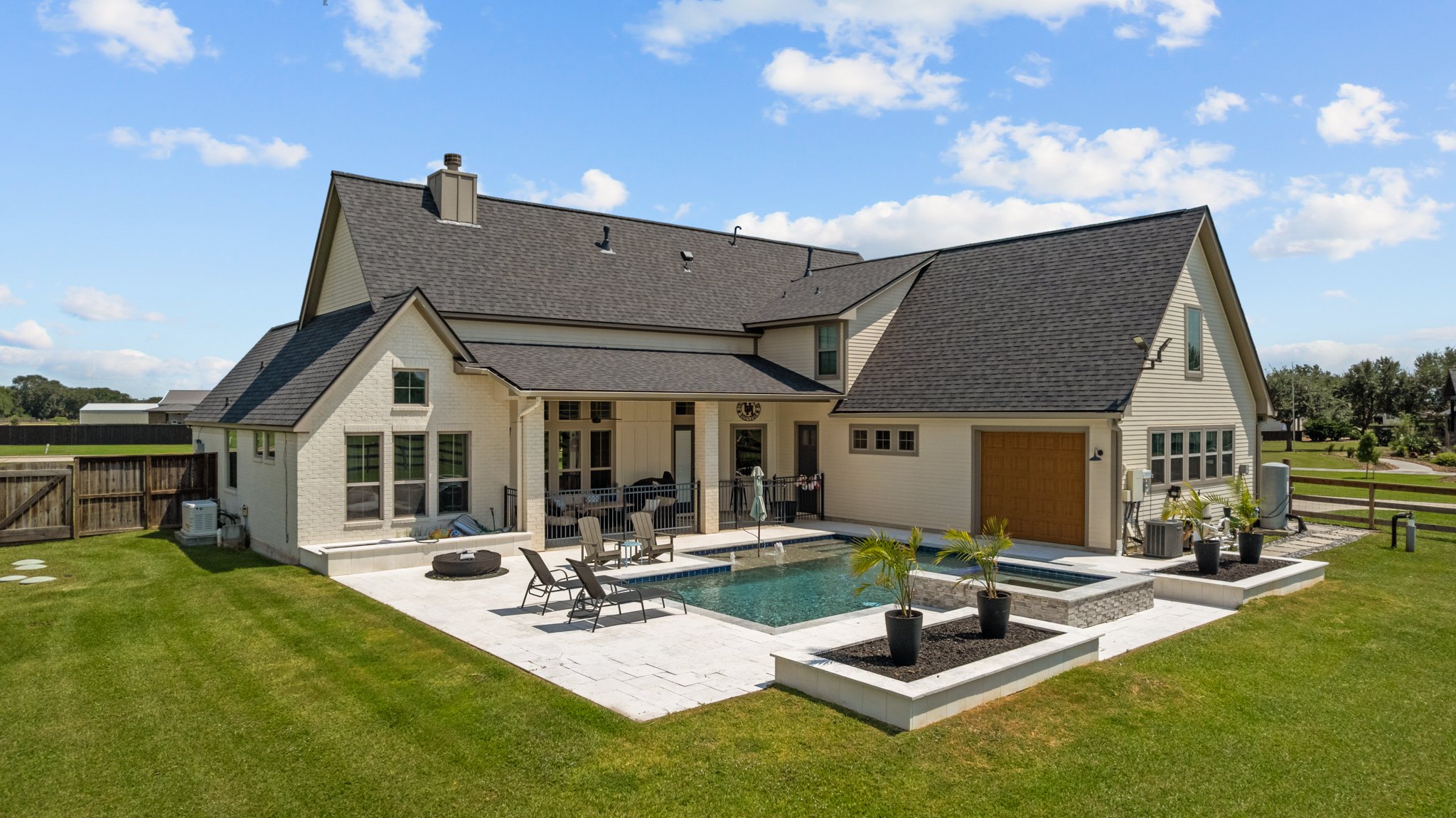 Modern two-story house with light-colored exterior, backyard with swimming pool, patio furniture, potted plants, and a well-maintained lawn, under a blue sky.