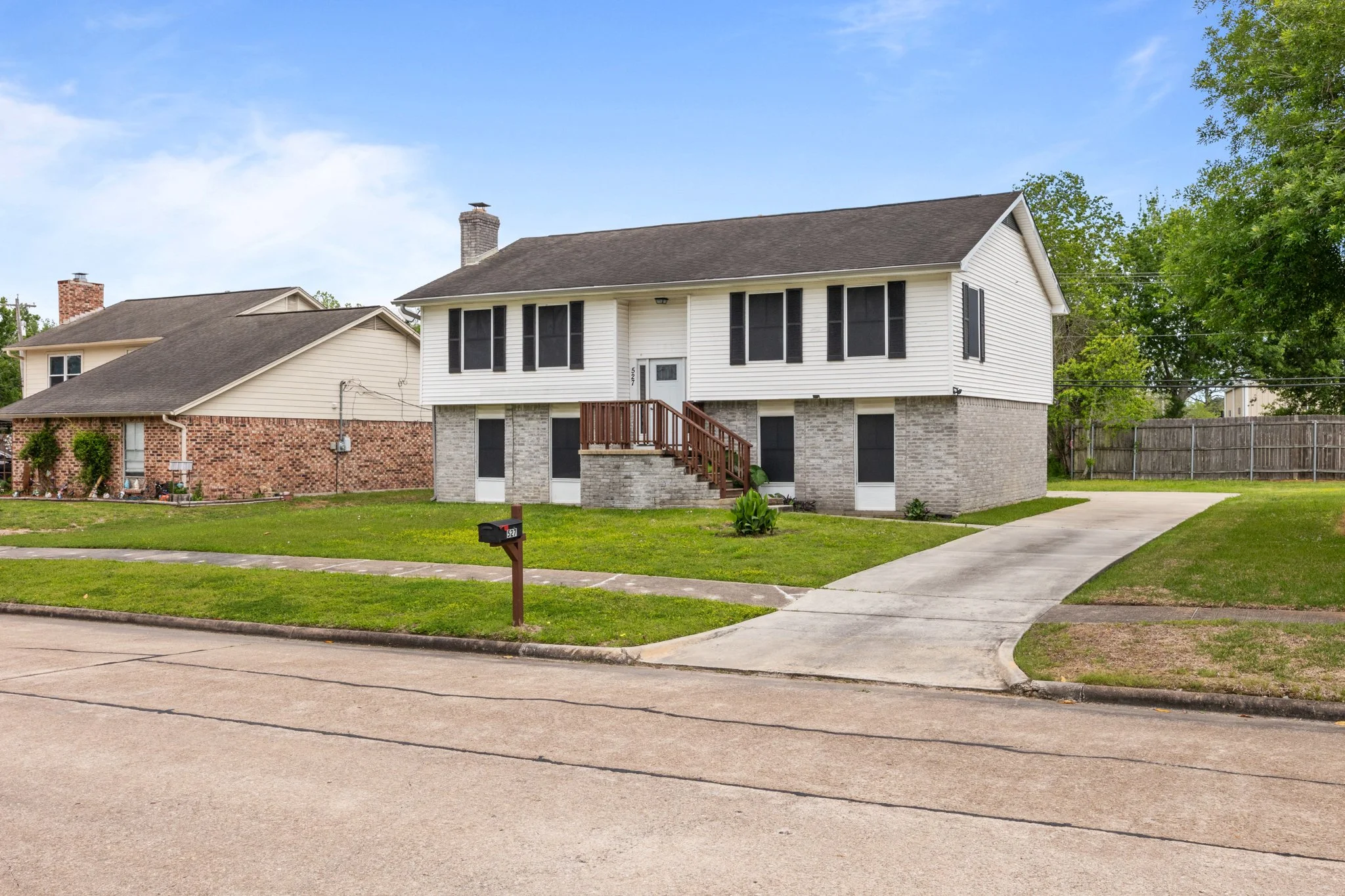 Front view of a two-story house with white siding, gray brick foundation, black shutters, a small front porch with stairs, a driveway on the right, green lawn, and a mailbox in the neighborhood with similar houses, trees, and a blue sky.