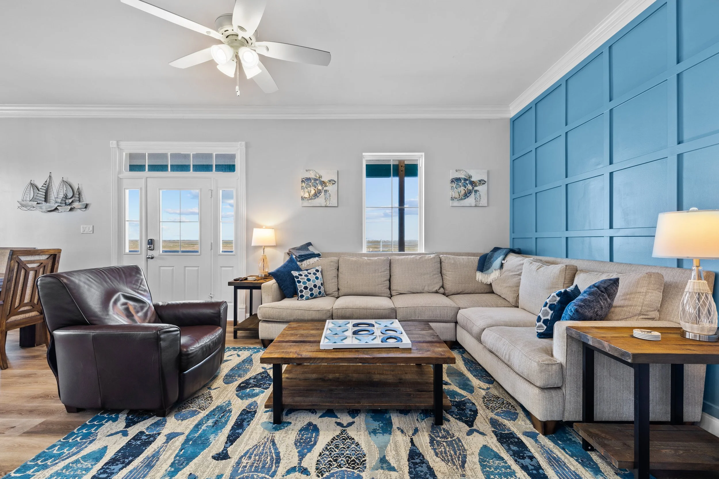 Living room with a beige sectional sofa, dark leather armchair, wooden coffee table, and blue wall with paneling. Decor includes ocean-themed artwork, lamps, and a patterned rug.