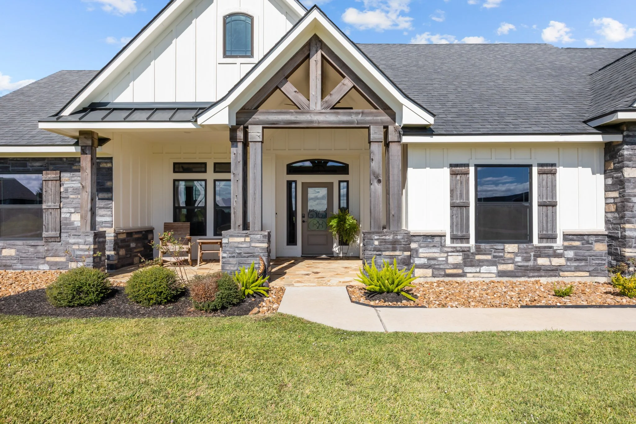Front view of a modern house with a landscaped lawn, stone accents, and wooden shutters, under a partly cloudy sky.