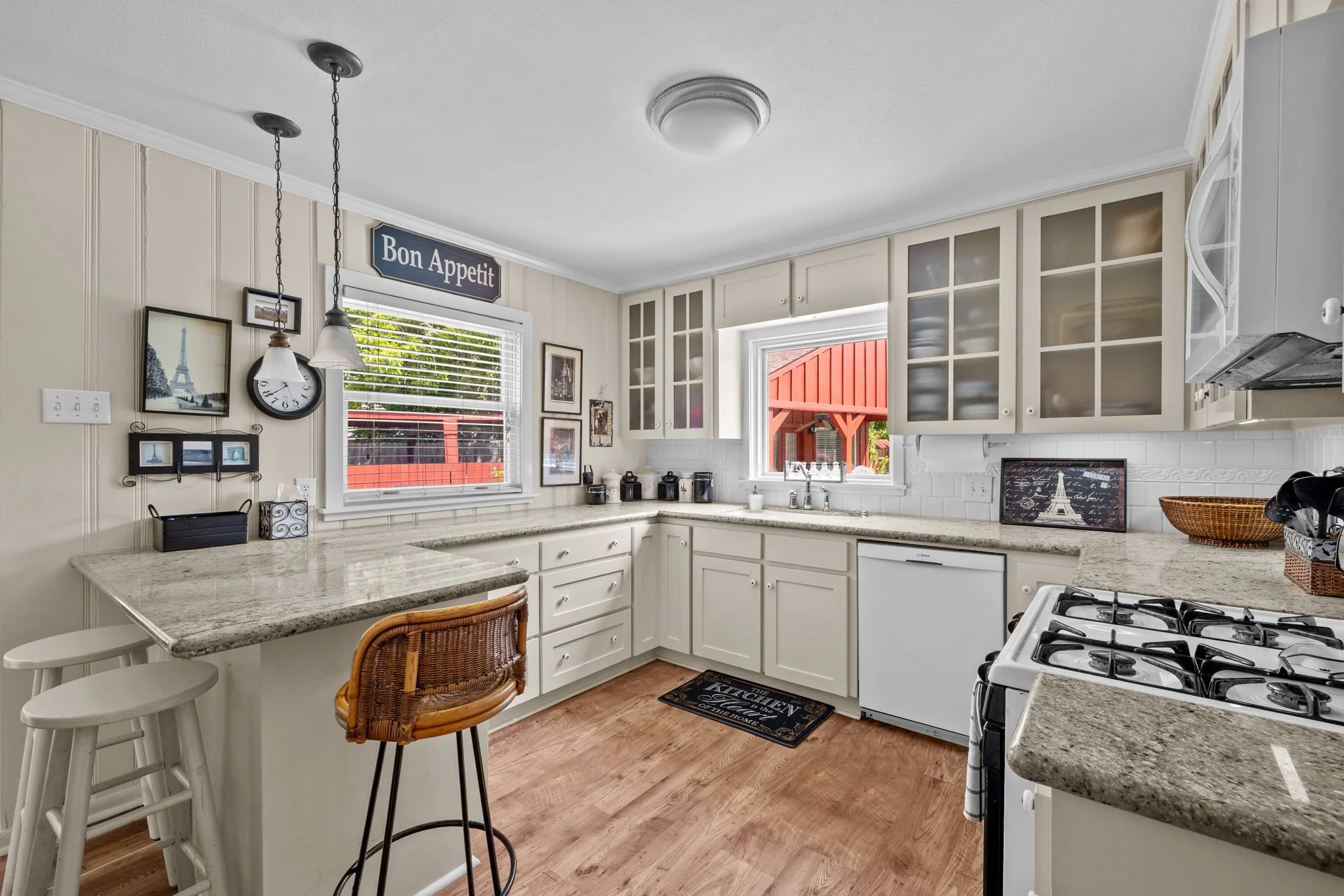 Kitchen with white cabinets, granite countertops, and a window with red exterior view. Decor includes a 'Bon Appetit' sign, Eiffel Tower pictures, and a Paris-themed framed art. There is a gas stove, dishwasher, and an island with two white stools and a wicker chair.