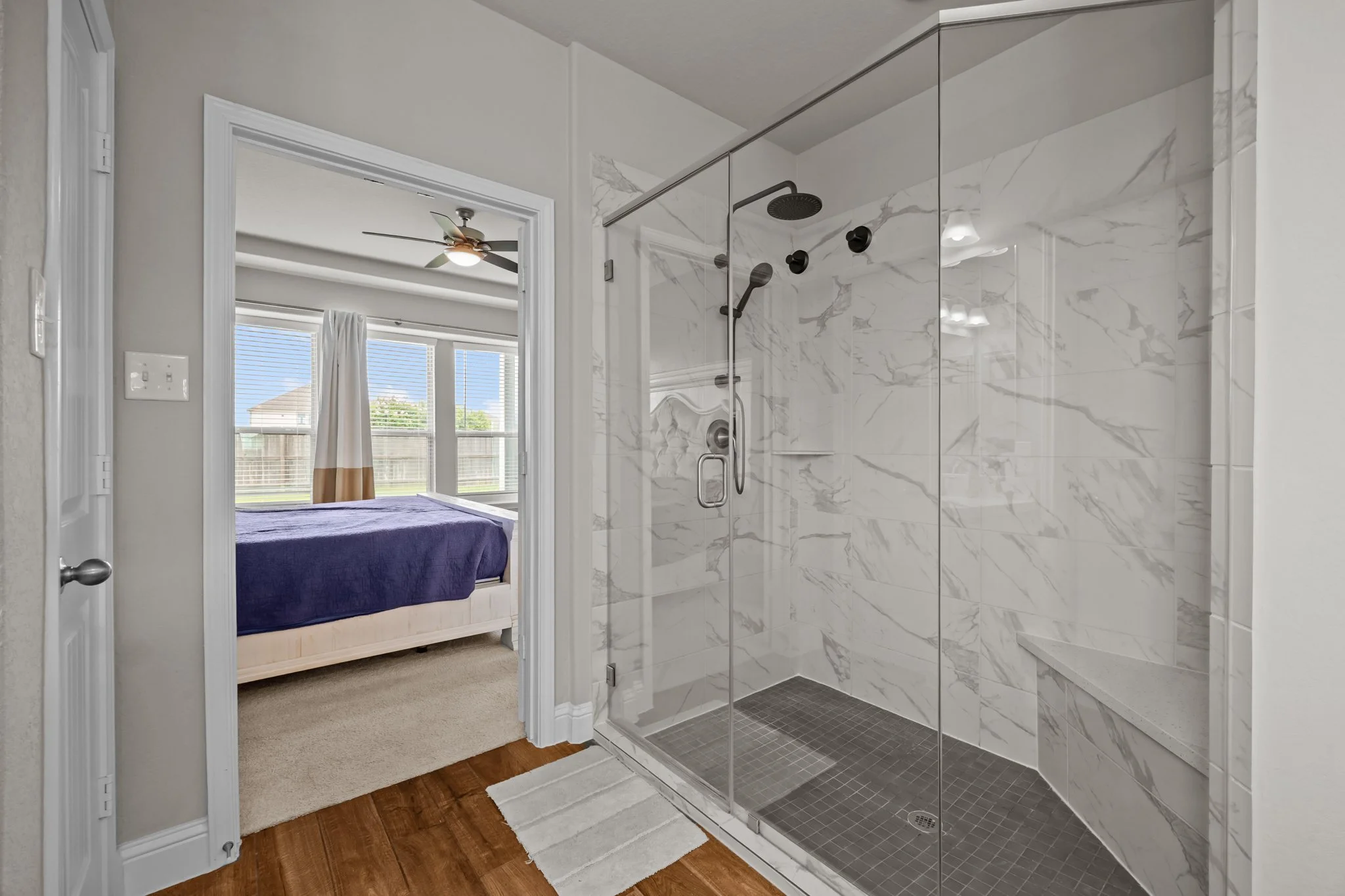 Modern bathroom with a glass-enclosed walk-in shower featuring white marble tiles with gray veins, a dark showerhead, and a bench, adjacent to a bedroom with a large window, beige carpet, and a bed with a dark blue blanket.