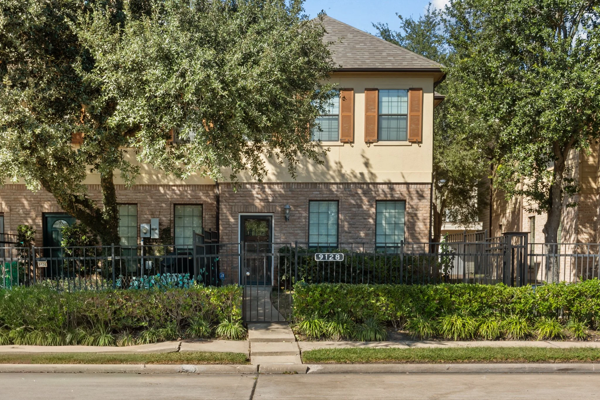 Front view of a two-story residential building with a brick and beige facade, surrounded by trees and green shrubbery, with a small sidewalk leading to the entrance.