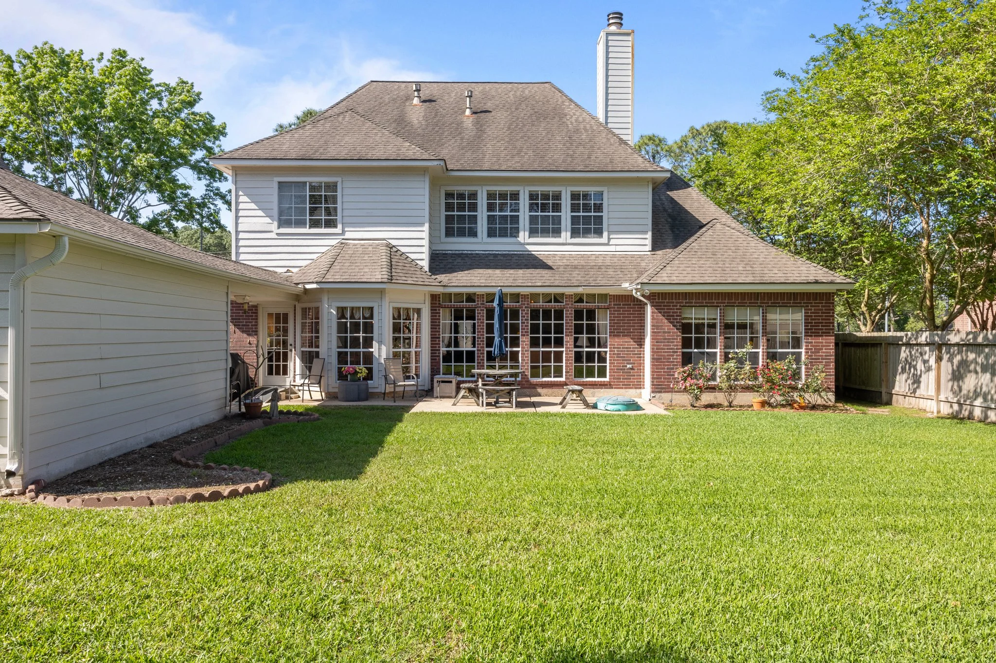 Backyard view of a two-story house with white siding and brick accents, large windows, a patio with outdoor furniture, a blue umbrella, and well-maintained green lawn with trees in the background.