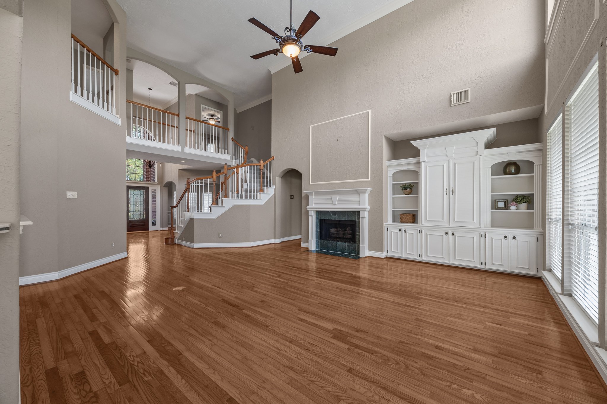 Empty living room with hardwood floors, white built-in shelves, a fireplace, large windows with blinds, and a ceiling fan.
