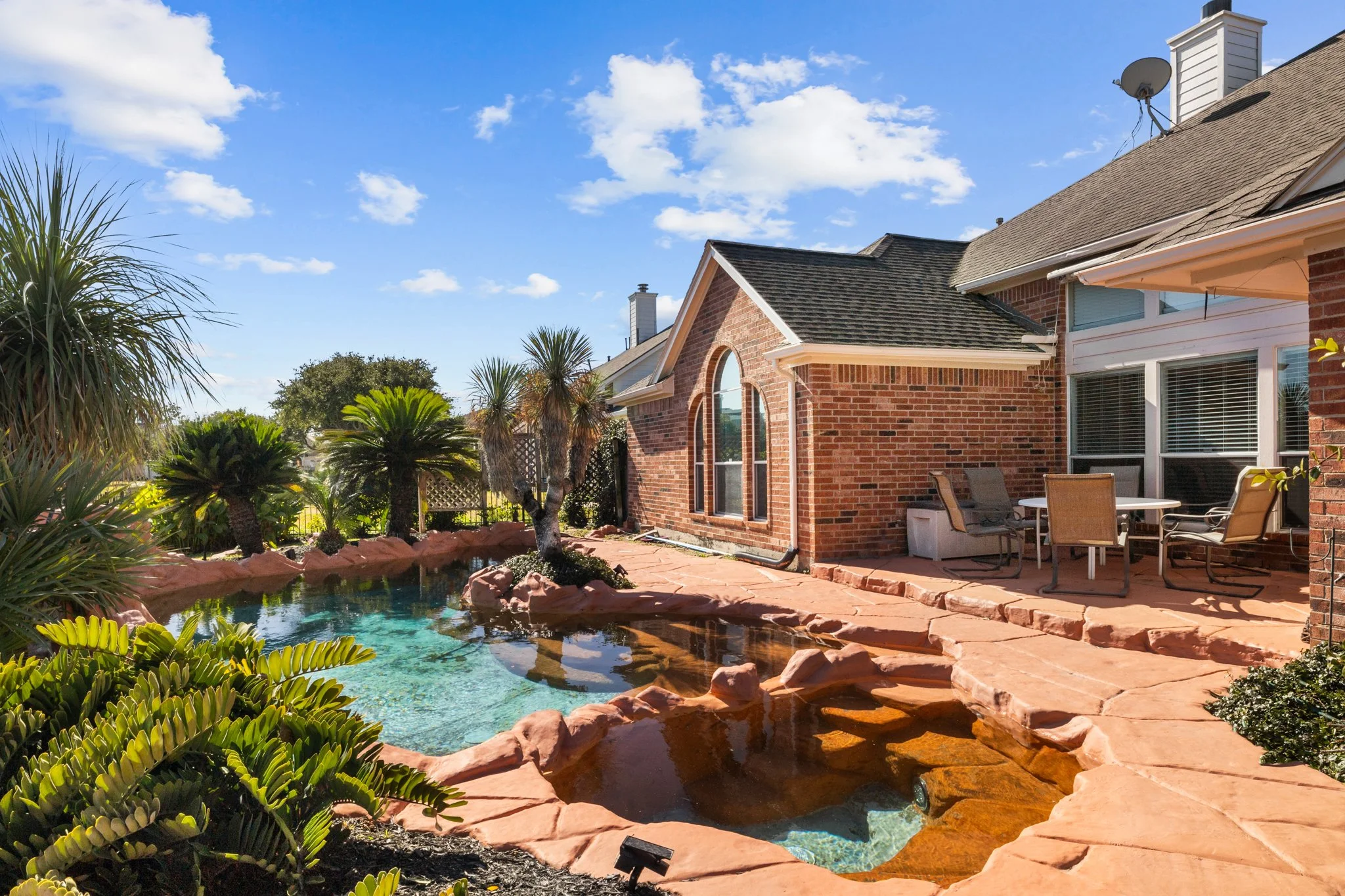 Backyard with a swimming pool, surrounded by desert plants and palm trees, adjacent to a brick house with patio furniture under a blue sky with clouds.
