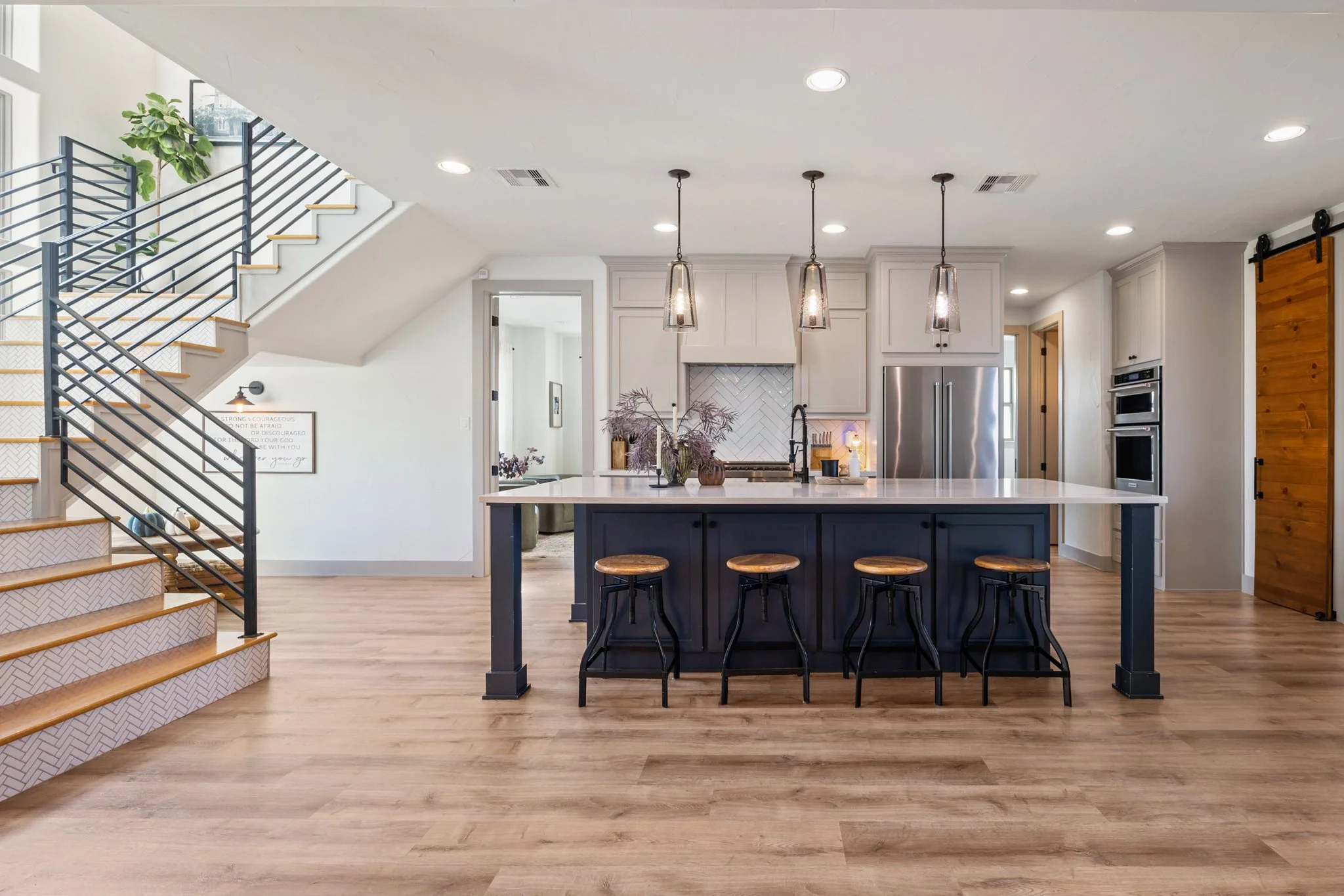 Modern kitchen with a large island, three barstools, white cabinetry, stainless steel appliances, wooden sliding barn door, and open-concept living space with stairs and hardwood floors.