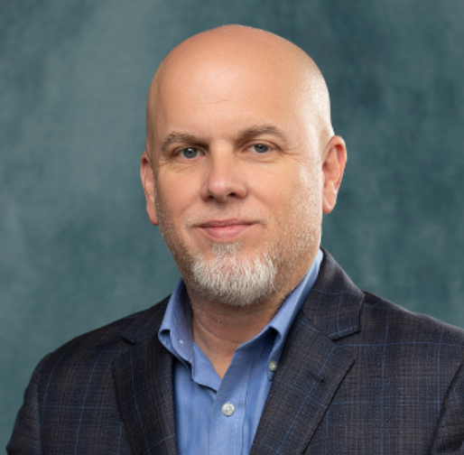 Close-up portrait of a bald man with a beard, wearing a blue collared shirt and dark blazer, against a textured teal background.