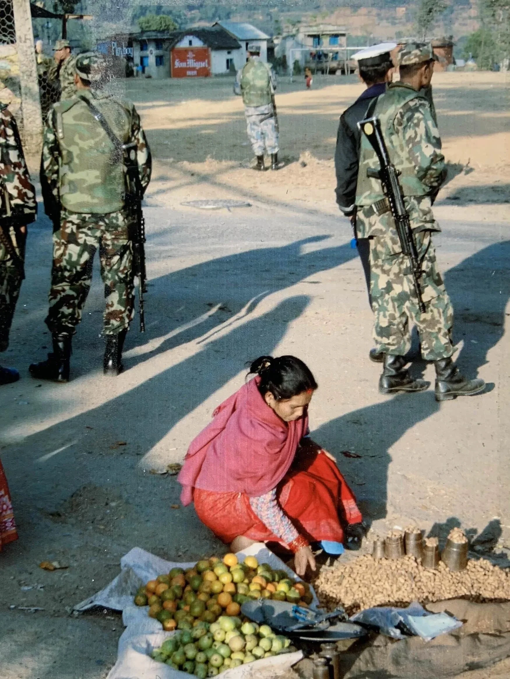 Nepali woman selling peanuts and limes, rifle-toting police in khaki at bus stop, Pharping, Nepal (~ 2006)  