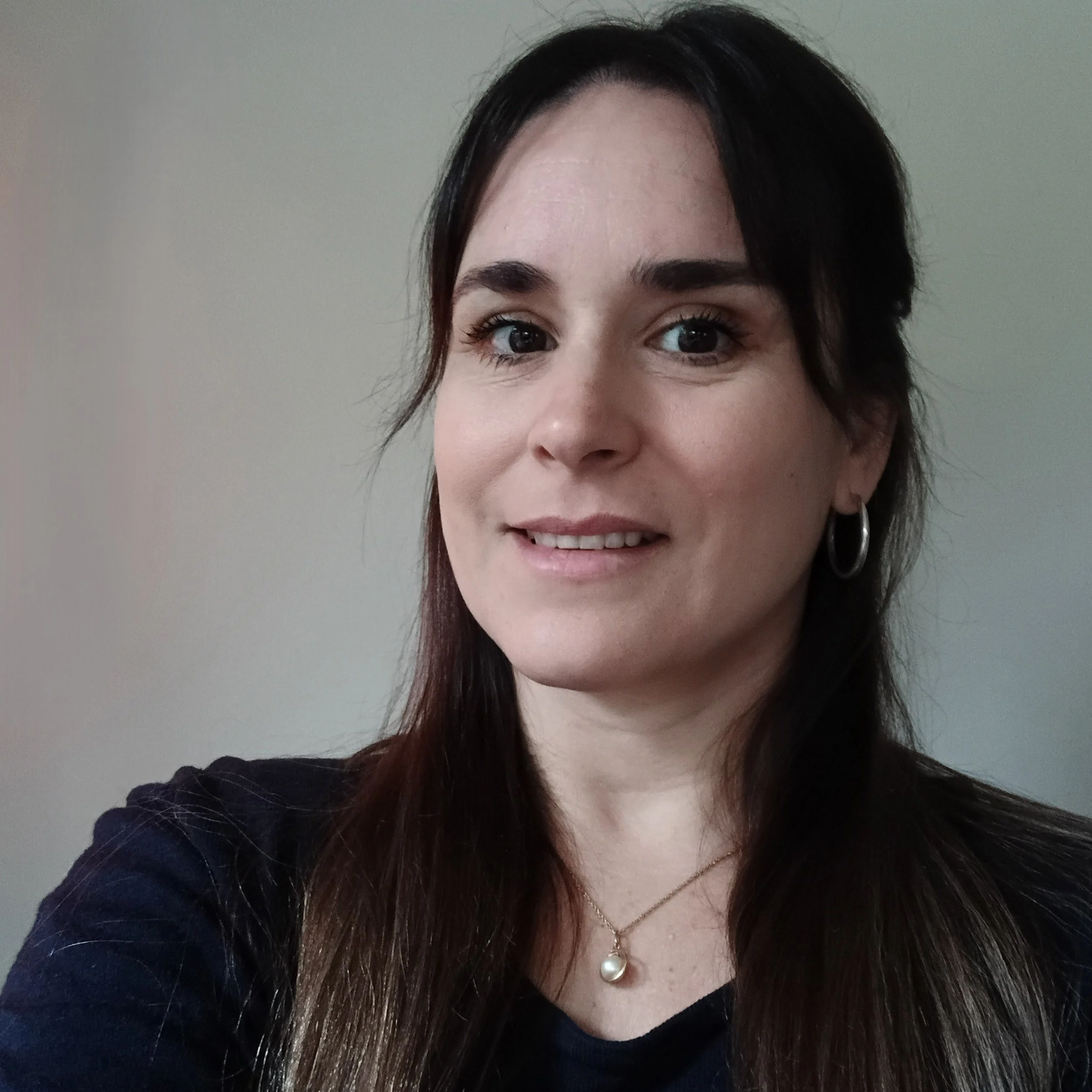 A woman with long dark hair, wearing hoop earrings and a pearl necklace, smiling at the camera against a plain background.