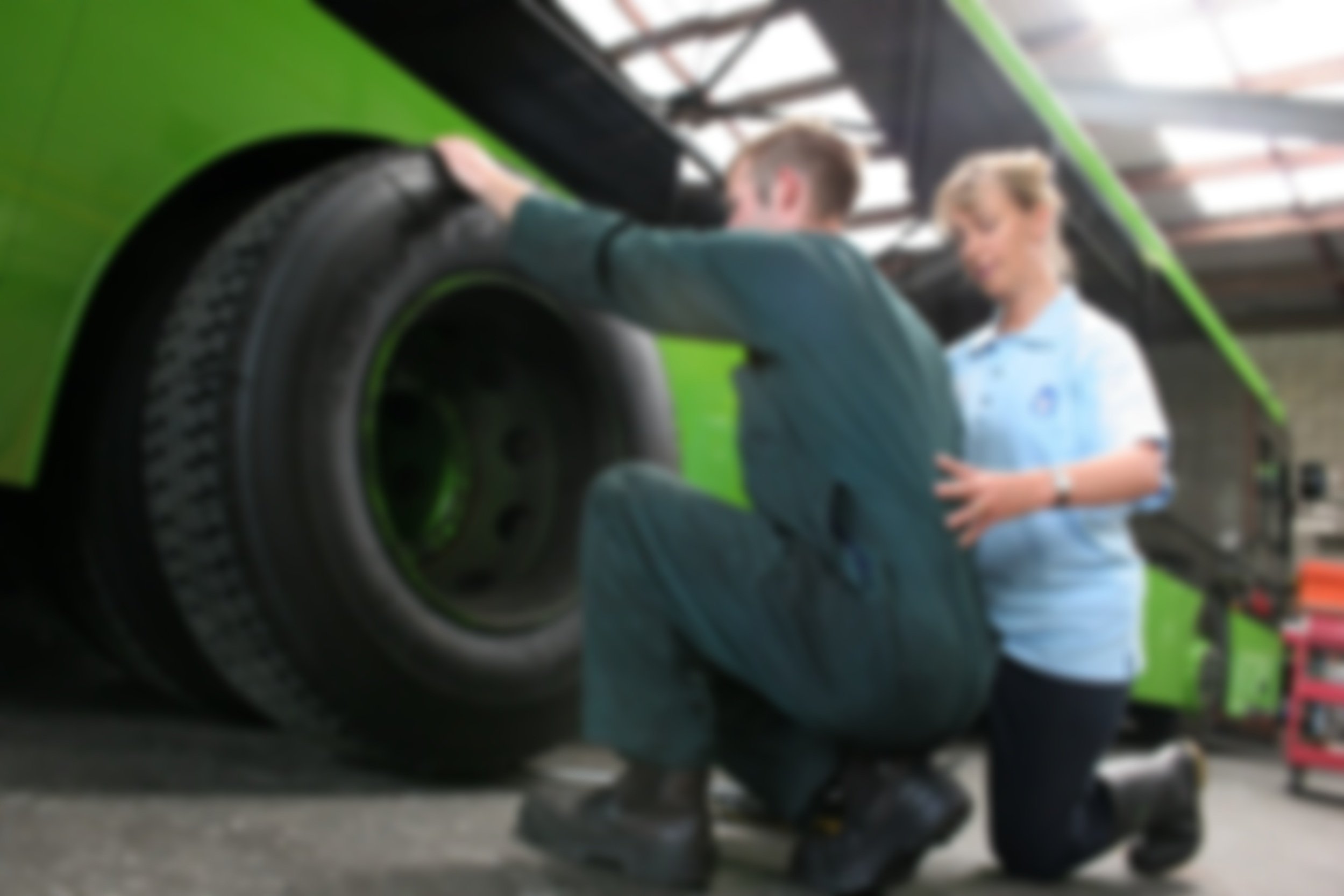 A person working on a large green bus tire while another person supervises in a garage or workshop.