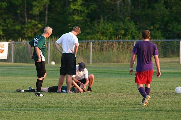 Four men on a soccer field with a ball, with one kneeling down and the others standing around, one approaching.