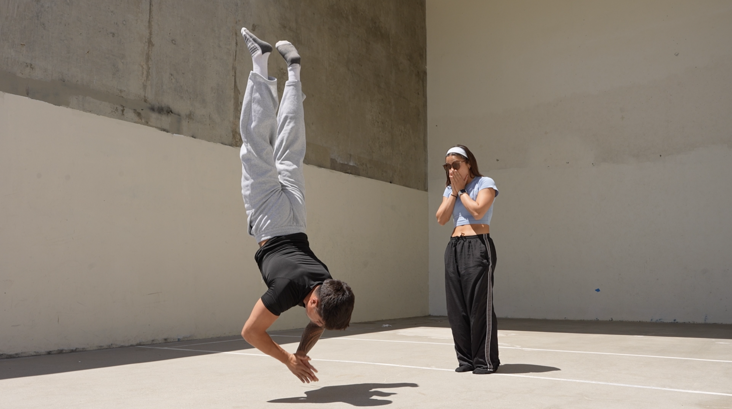 A person is performing a handstand on a basketball court, while another person is standing nearby with hands covering their mouth, watching. The person doing the handstand is upside down with legs straight up, wearing gray sweatpants, a black shirt, and white socks. The observer is wearing sunglasses, a white headband, a light blue crop top, and dark athletic pants.