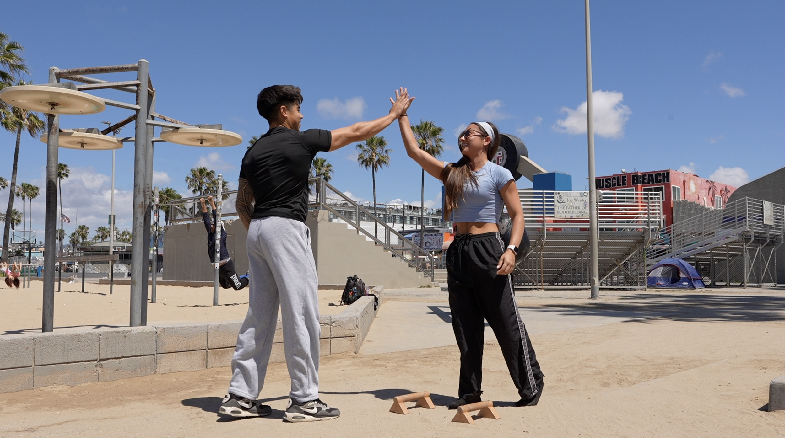 Two people practicing high fives on a sunlit outdoor beach volleyball court, with equipment, a tent, and buildings in the background.