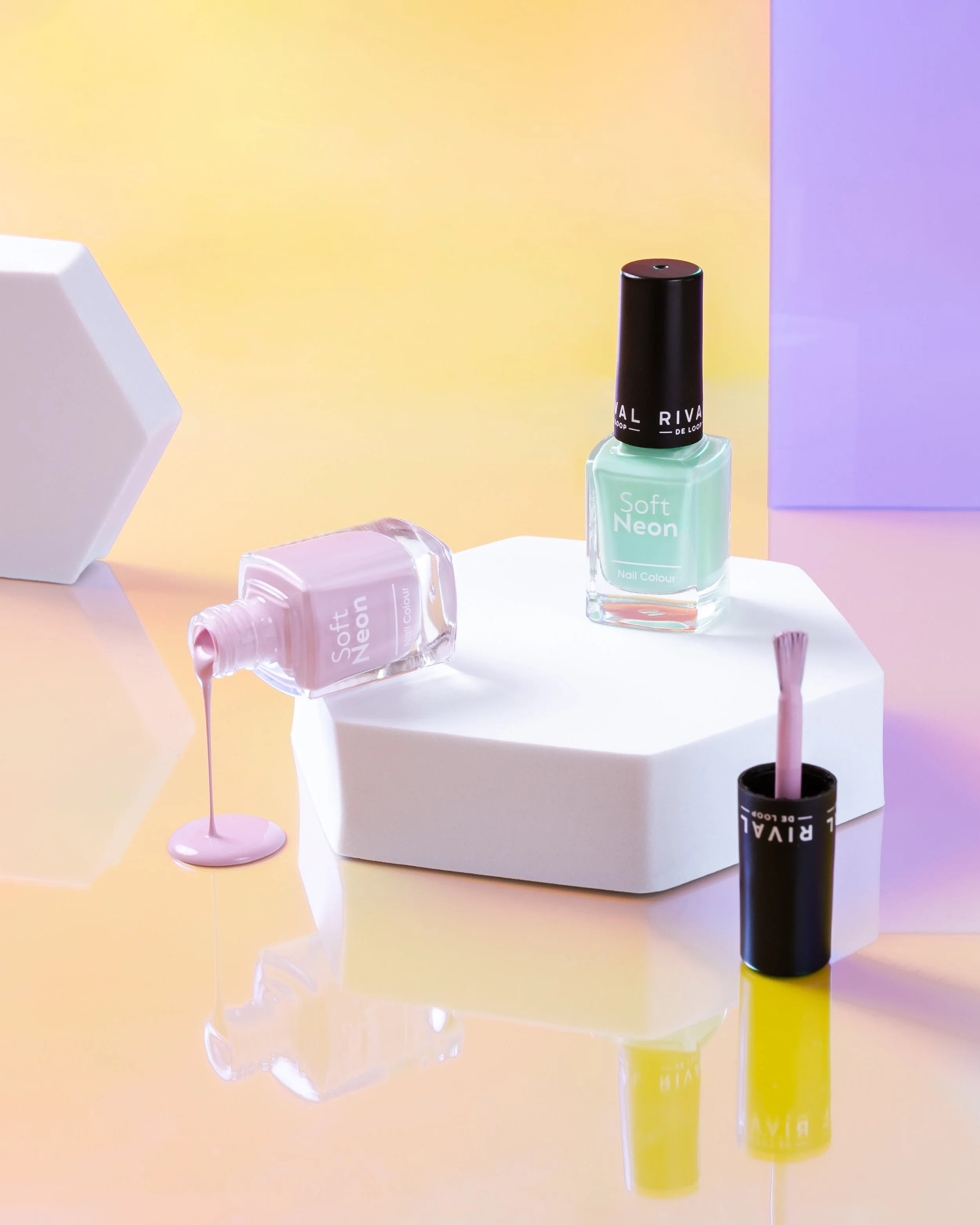 Zwei Nagellackflaschen mit der Bezeichnung "Soft Neon" auf einem weißen Plattform. Eine Flasche hat rosa Lack und ist umgekippt, wobei der Nagellack ausläuft. Die andere Flasche ist mintgrün und steht aufrecht. Im Hintergrund ist ein bunter, pastellf