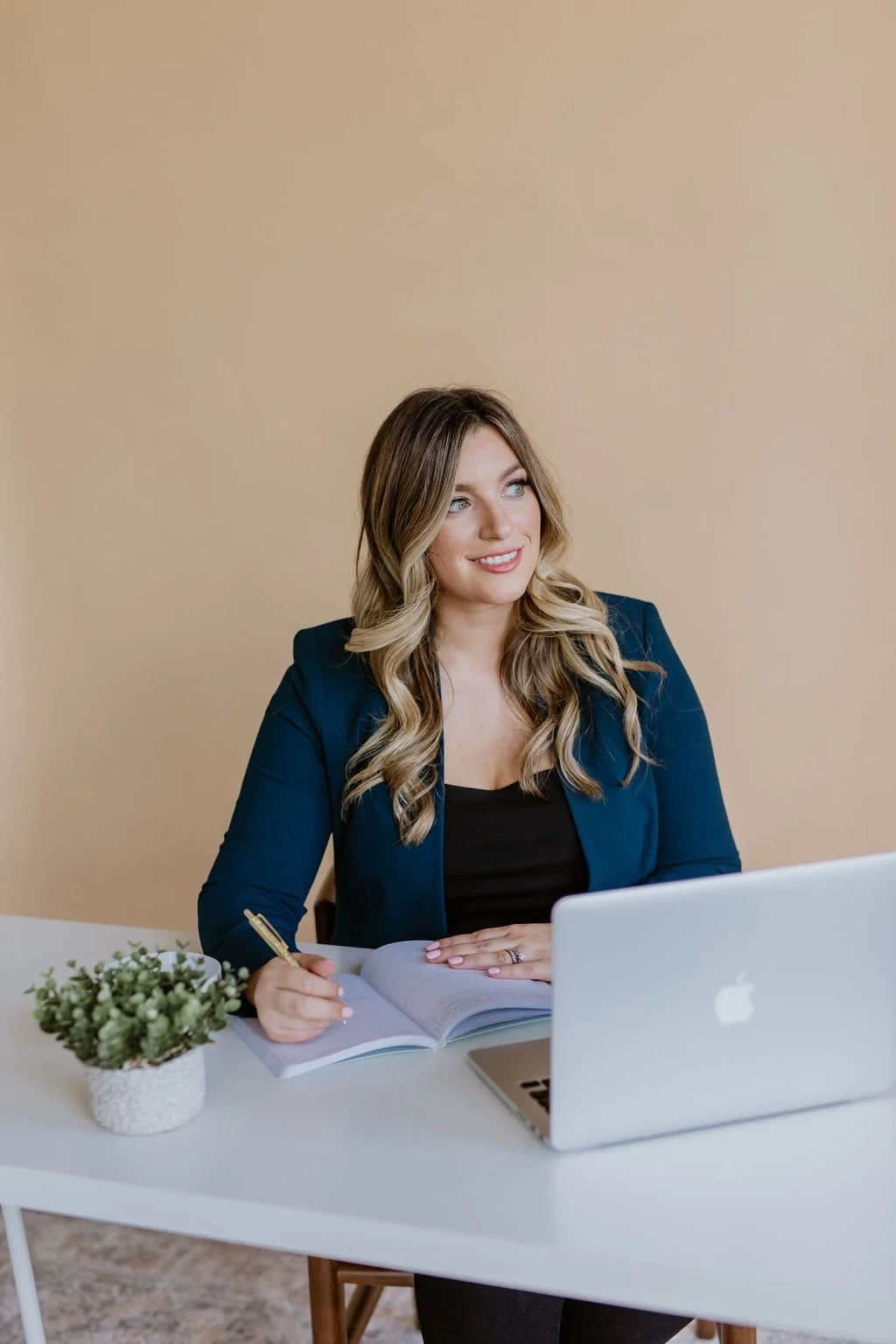 A woman with long wavy blonde hair, wearing a blue blazer and black top, sitting at a white desk with a silver laptop, a notebook, a pen, and a small plant with green leaves, looking to the side with a smile.