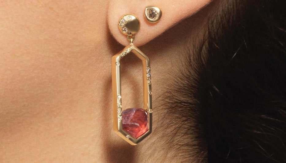 Close-up of a person's ear wearing two gold earrings, one stud and one geometric with a red stone.